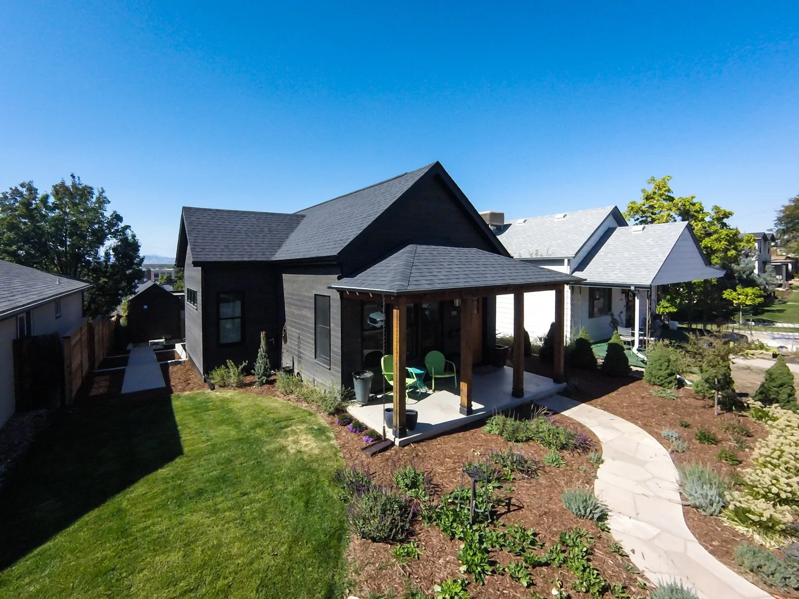 A modern house with black and white exterior, a small porch with green chairs, surrounded by a landscaped yard with a curved stone pathway, bushes, and trees under a clear blue sky. Roofing Company in Denver Colorado