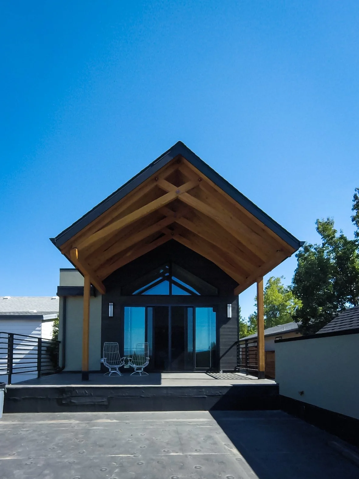 Modern house with a large wooden front porch and roof, glass sliding doors, two outdoor chairs, and a landscaped driveway. Roofing Company in Denver Colorado