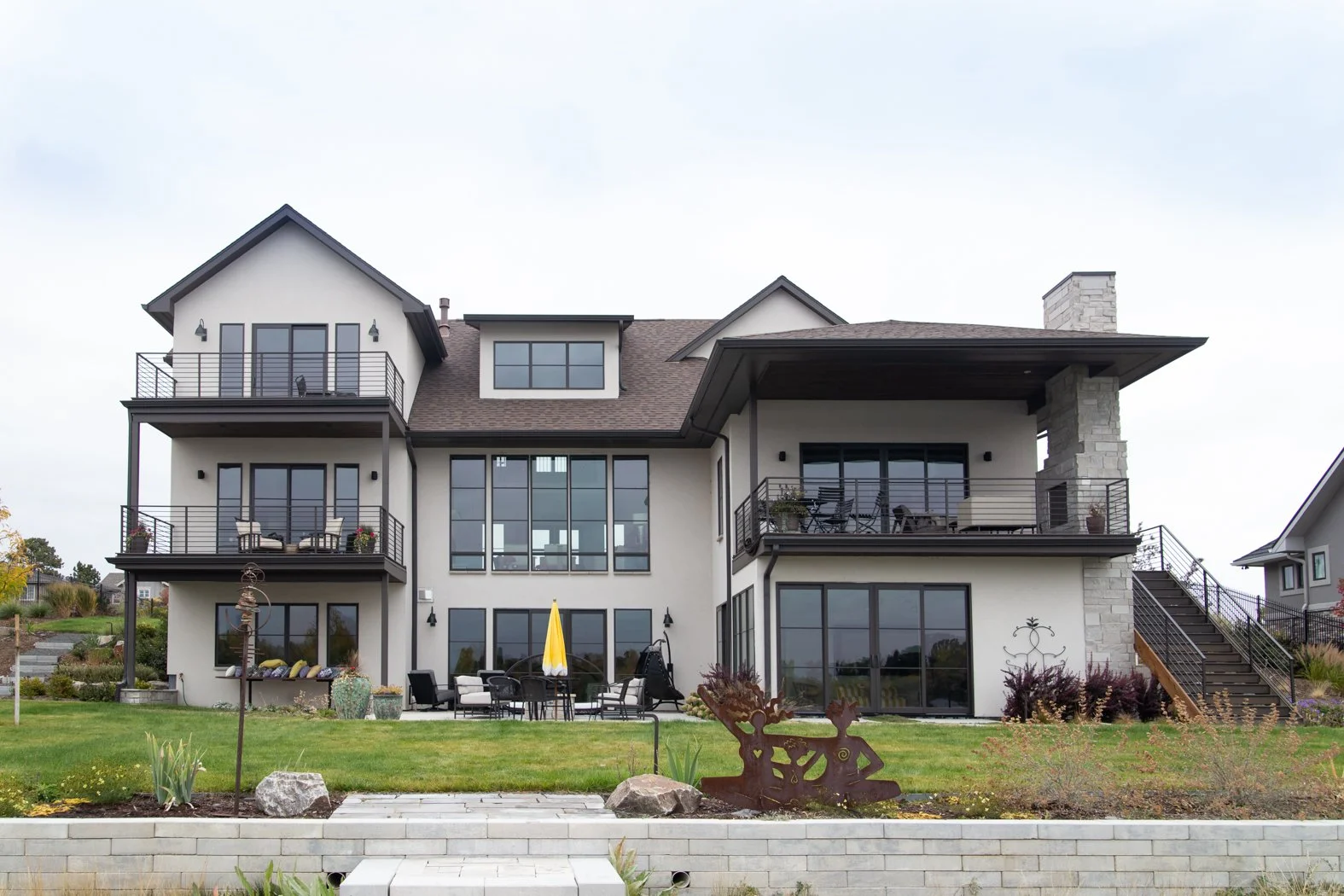 Modern multi-story house with multiple balconies and large windows, surrounded by a landscaped yard with outdoor furniture, plants, and decorative sculptures. Roofing Company in Denver Colorado