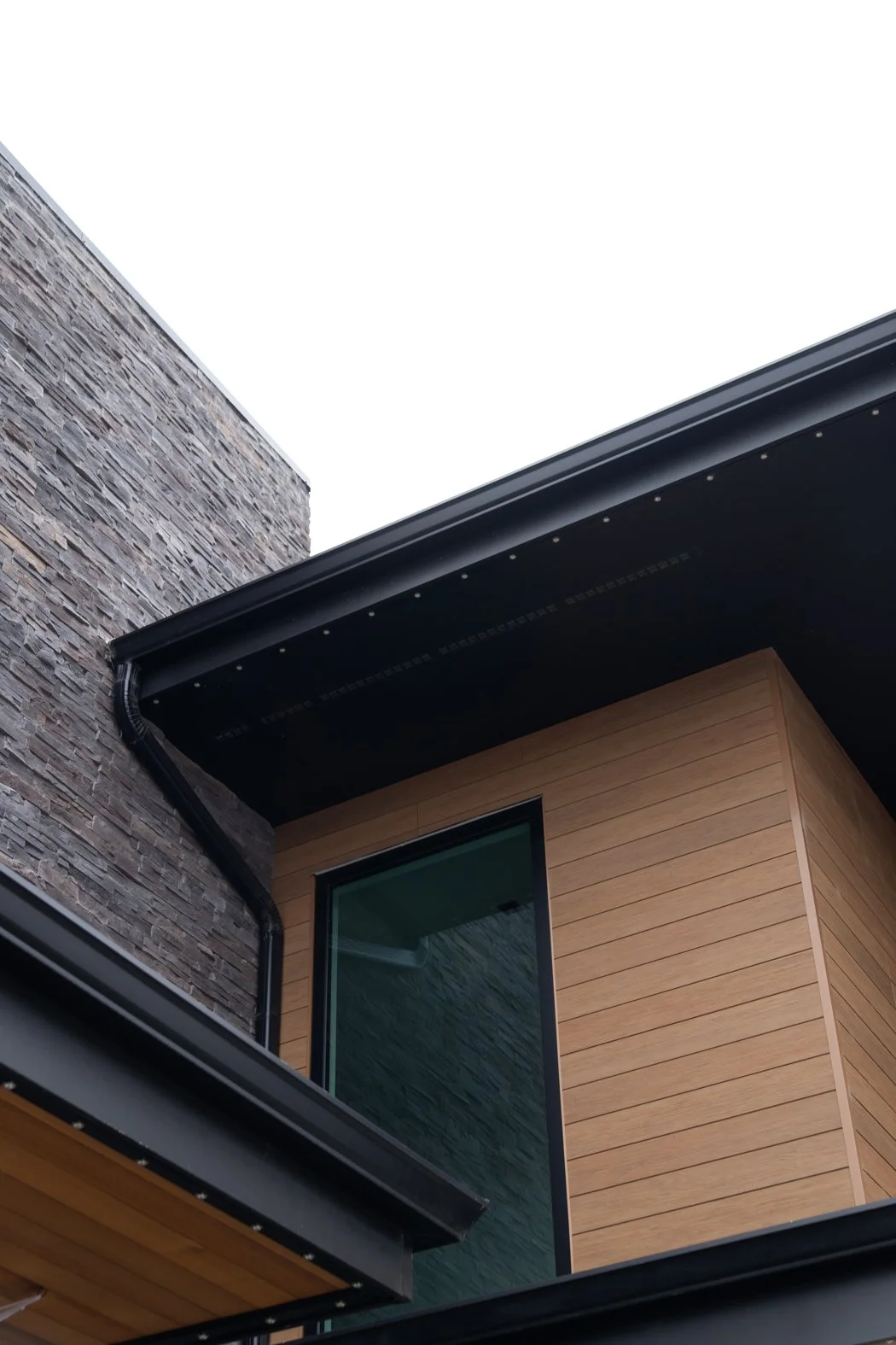 Close-up view of a modern residential building exterior featuring a stone wall, wooden siding, a large window, and a black roof overhang. Roofing Company in Denver Colorado