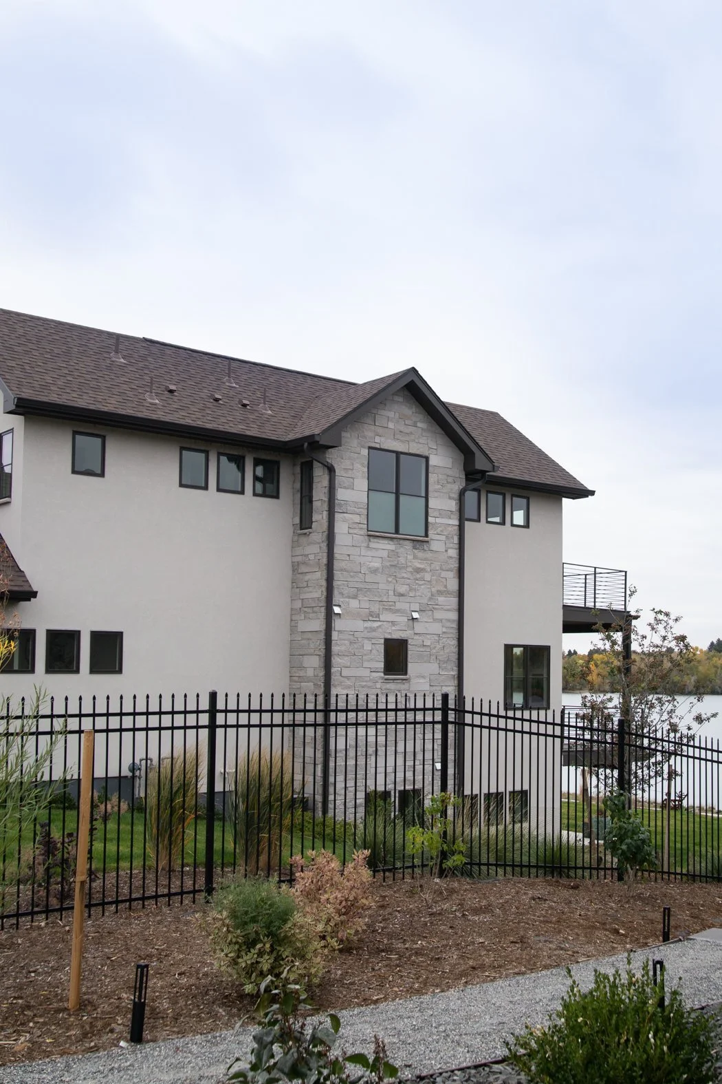 A modern multi-story house with gray stone and beige siding exterior, black window frames, and a brown sloped roof, situated near a water body, with a black metal fence and landscaped garden area in the foreground. Roofing Company in Denver Colorado