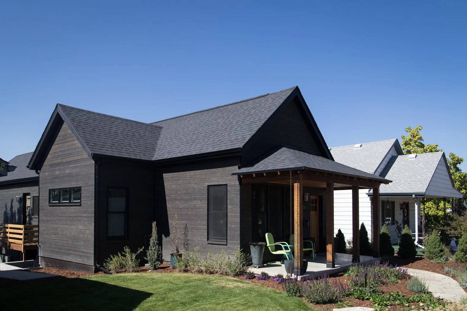 Exterior view of a modern black house with a small front porch, green patio chairs, and landscaped lawn under a clear blue sky. Roofing Company in Denver Colorado