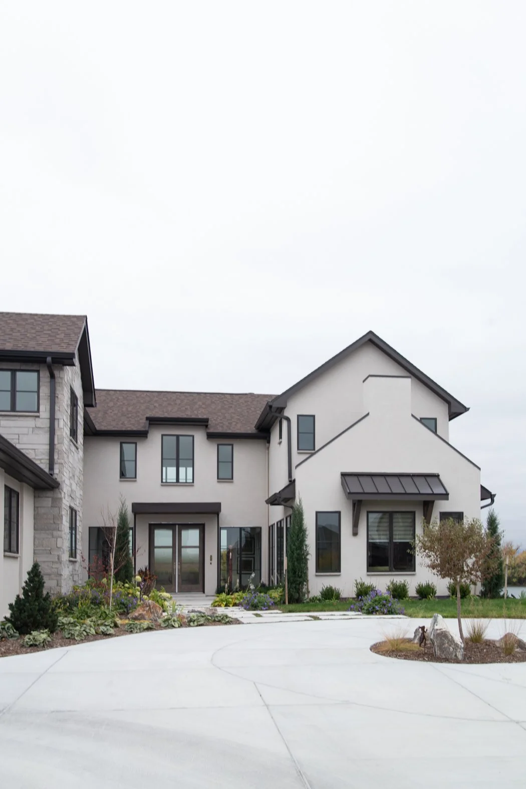 front view of a modern multi-story house with white exterior walls, black window frames, and a brown shingled roof, with landscaped garden and driveway in the foreground. Roofing Company in Denver Colorado