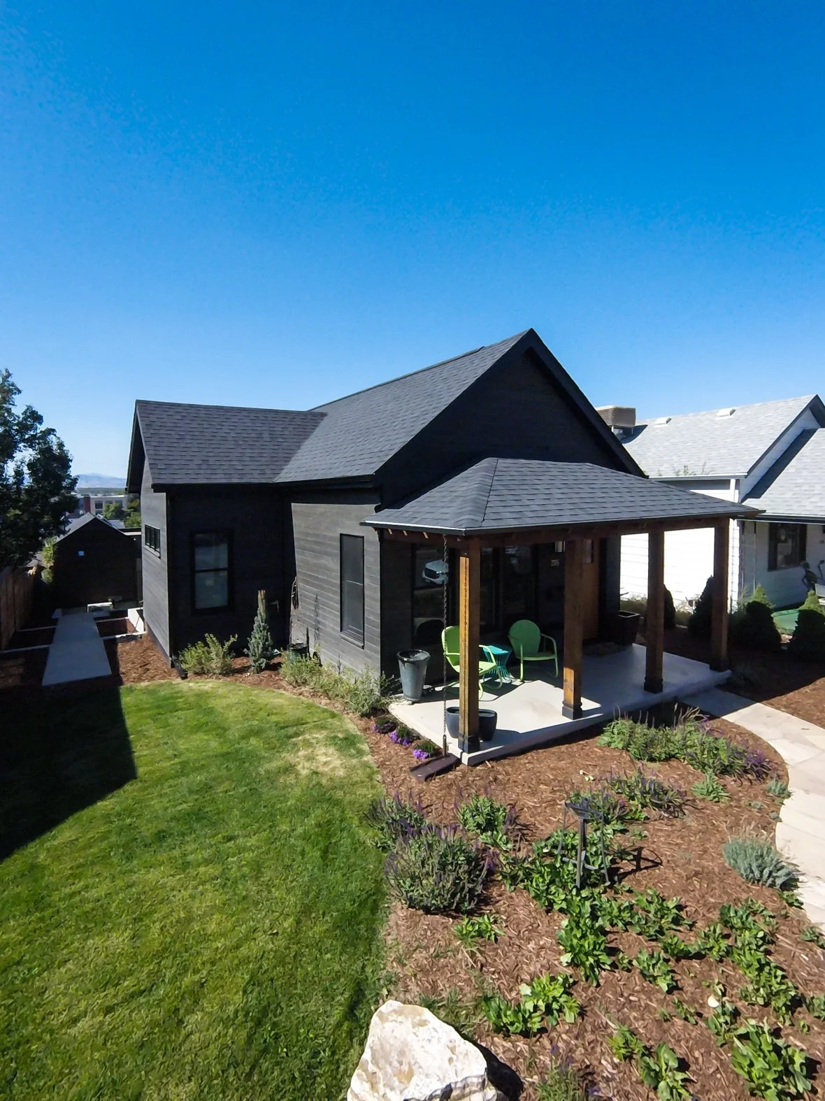 A backyard with a small dark gray house featuring a covered porch with seating, surrounded by a well-maintained lawn, flower beds, and a concrete pathway, under a clear blue sky. Roofing Company in Denver Colorado
