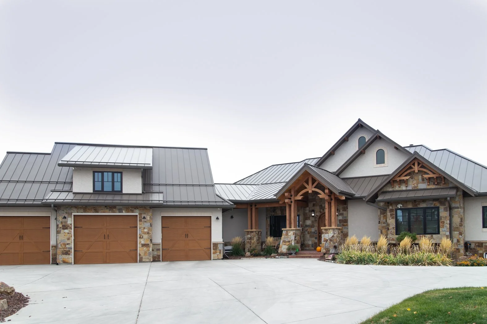 A large suburban house with a stone and stucco exterior, metal roof, three garage doors, and a landscaped front yard with ornamental grasses and pumpkins. Roofing Company in Denver Colorado