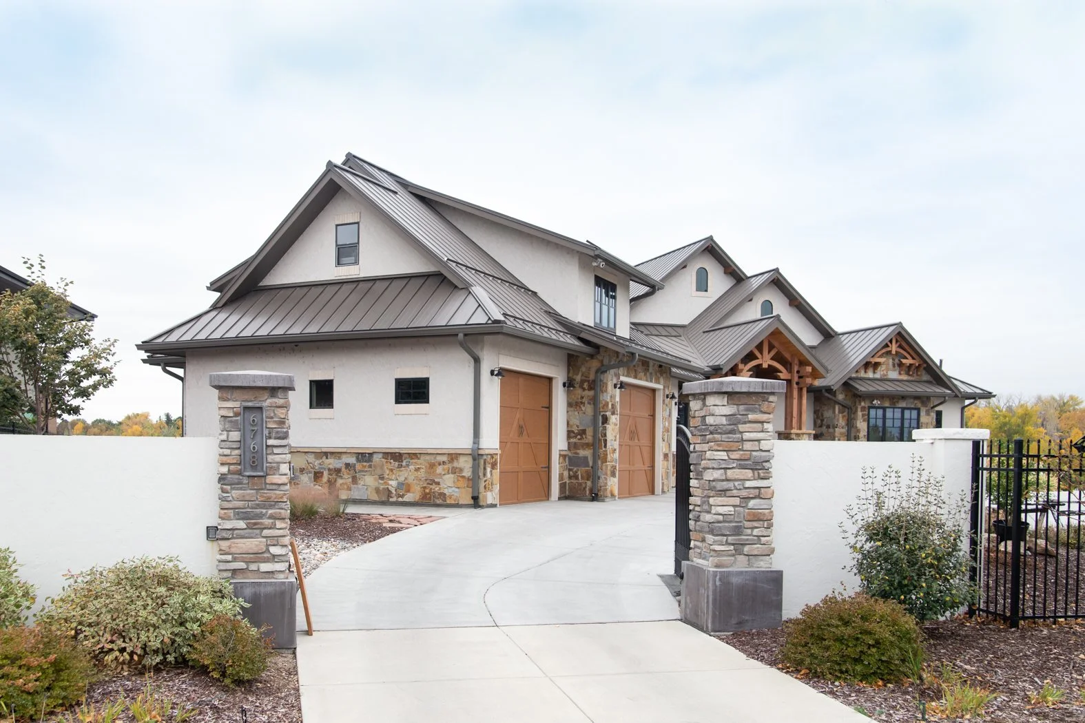 Modern house with stone and stucco exterior, metal roof, and front gate flanked by stone pillars, surrounded by landscaped yard. Roofing Company in Denver Colorado