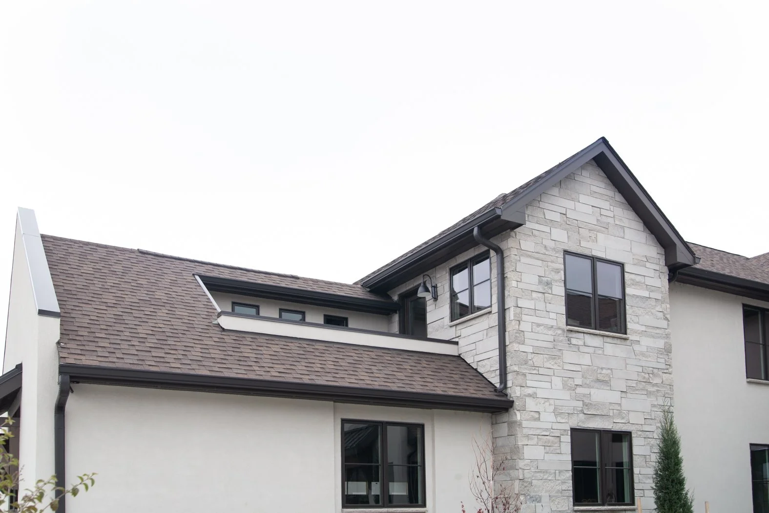 Modern house exterior with stone and stucco finish, black framed windows, and a pitched roof with brown shingles. Roofing Company in Denver Colorado