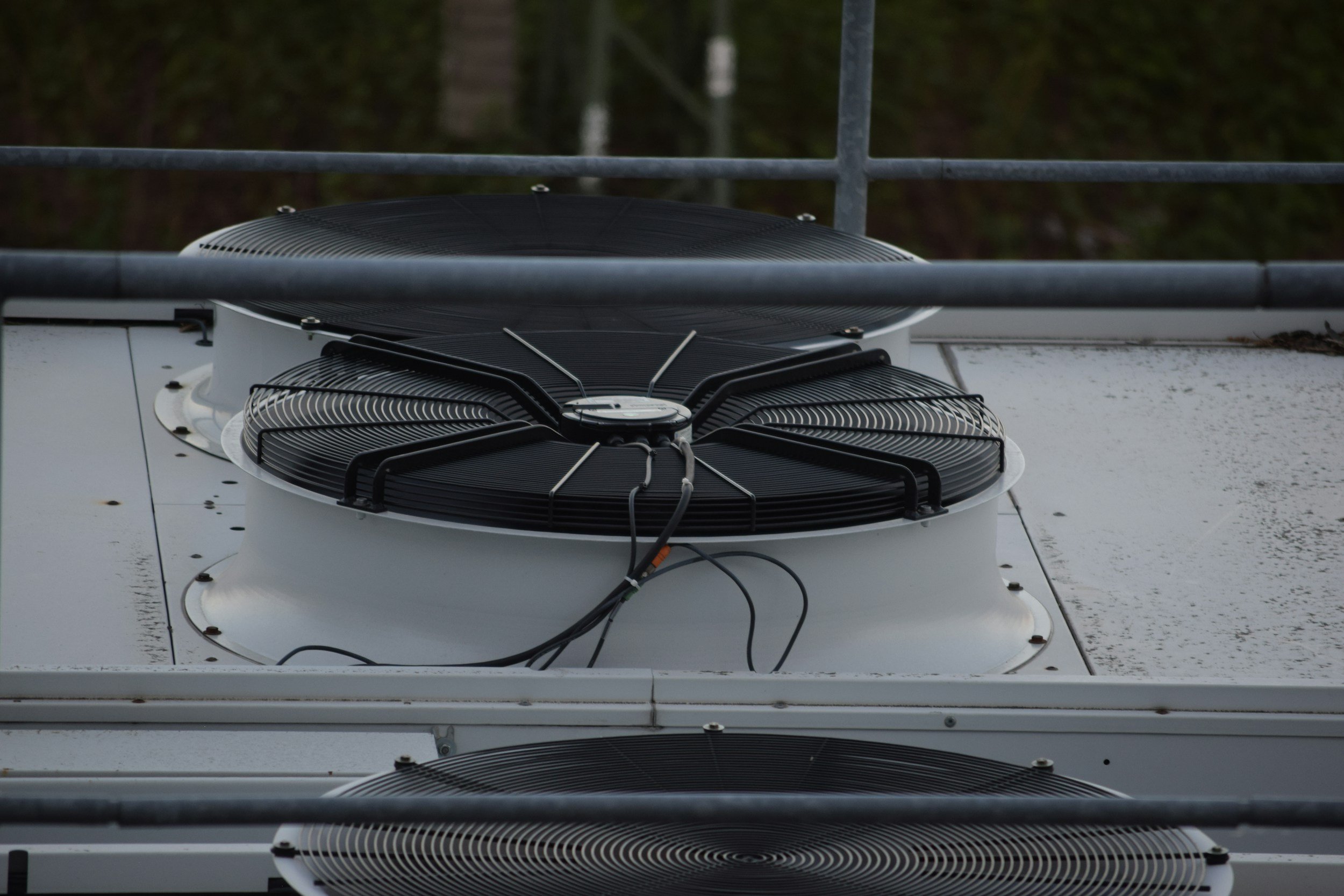 Close-up view of rooftop HVAC or cooling system units with black fans and connected wires on a flat white rooftop, surrounded by safety railings.