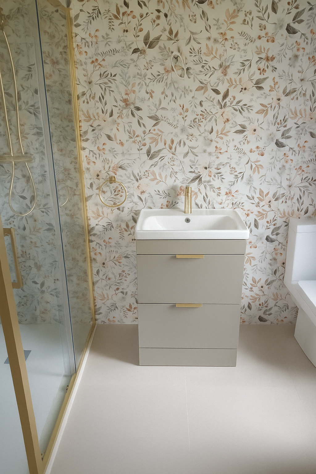 A bathroom with floral patterned wallpaper, a white sink with a gold faucet and handles, a small cabinet below the sink, a white toilet to the right, and a glass shower enclosure with gold trim on the left.