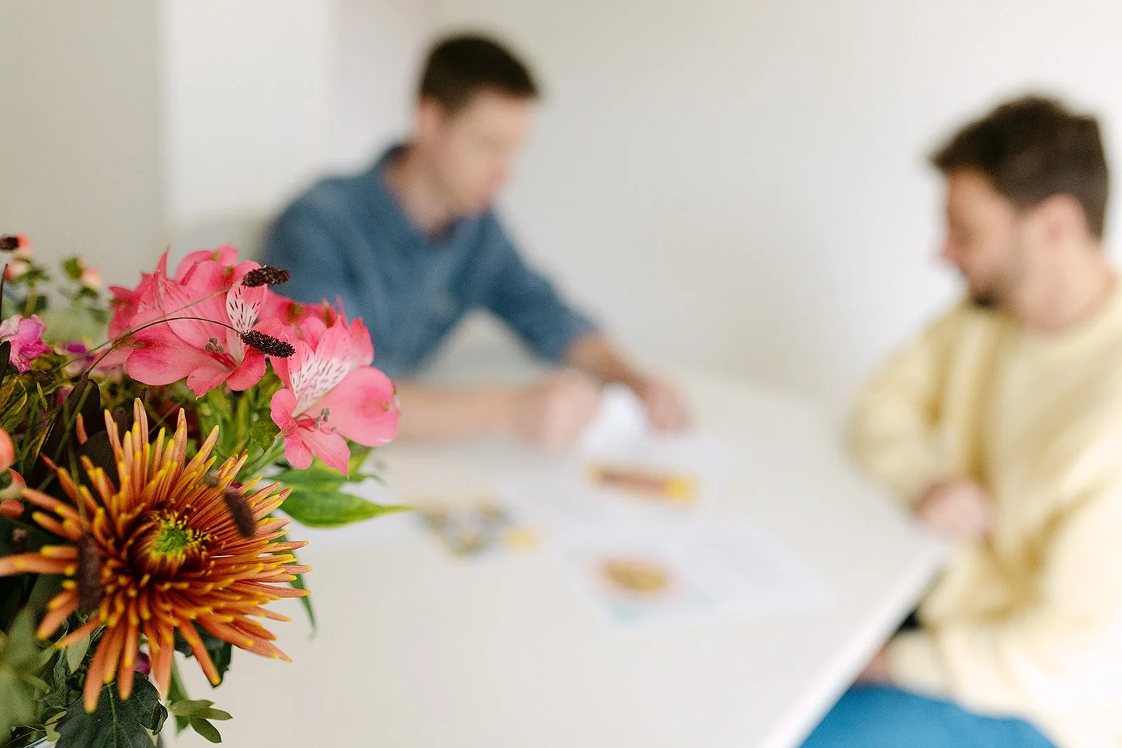 Close-up van een boeket bloemen, inclusief roze lotussen en een oranje-bruine bloem, met twee mensen op de achtergrond die aan tafel zitten, maar ze zijn onscherp.