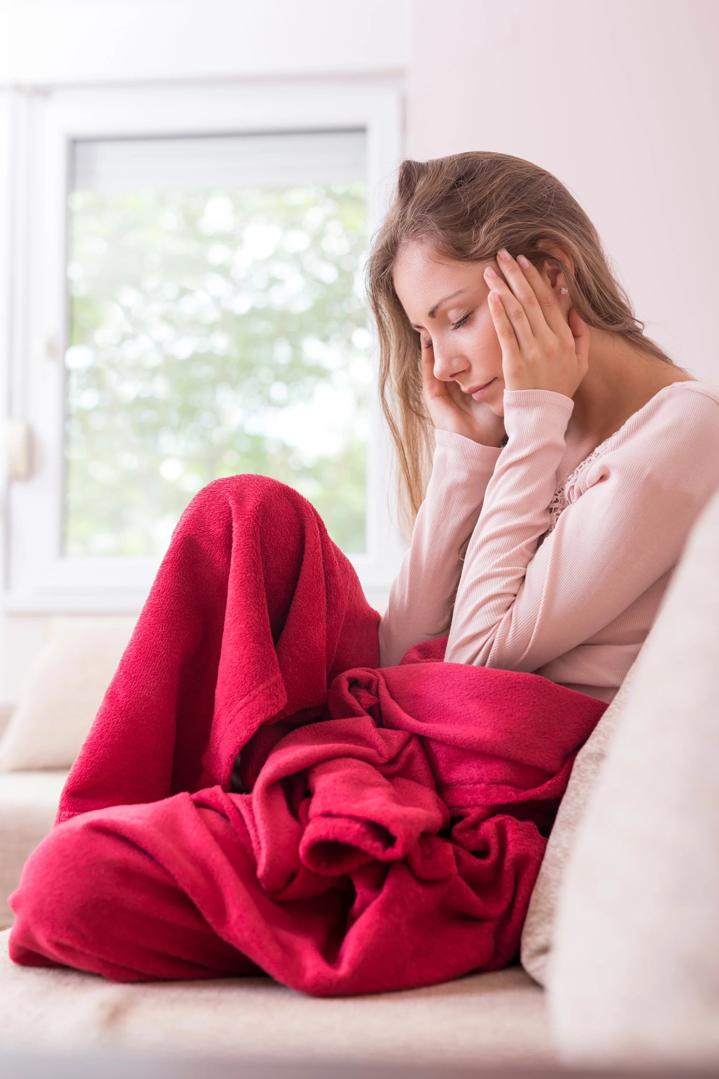Vrouw zit op bank met hoofdpijn, omringd door een rood dekentje, kleurrijk en comfortabel interieur, raam met uitzicht op groene bomen.