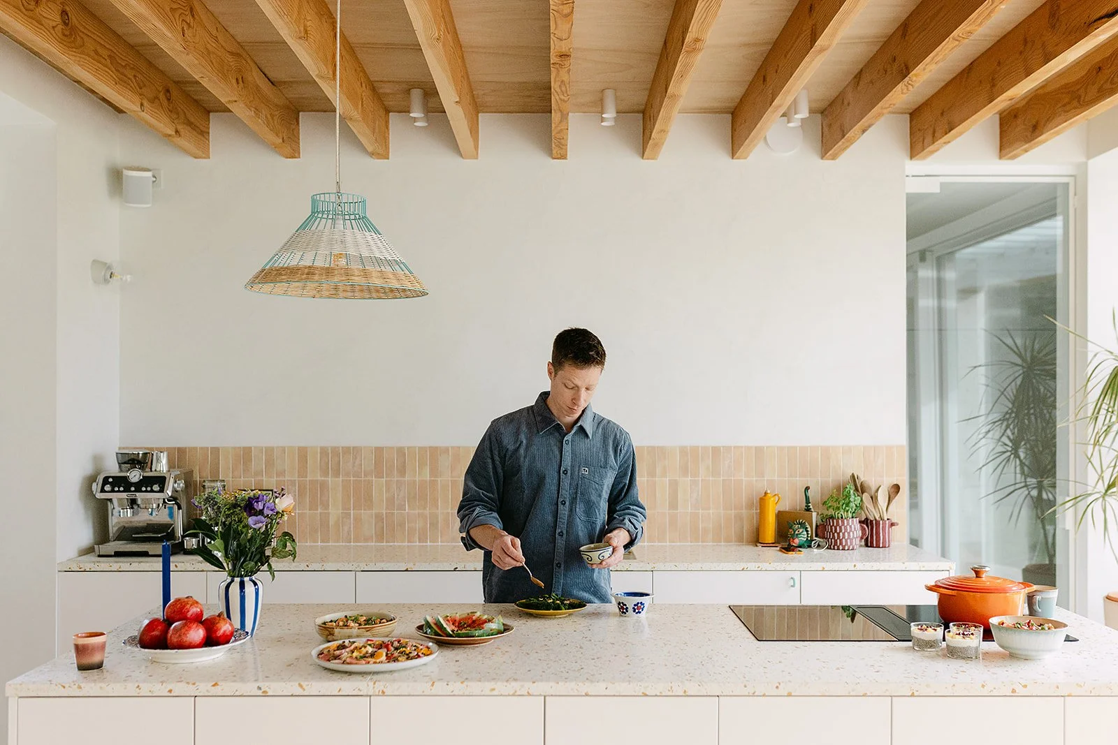 Man staat in een lichte, moderne keuken en serveert eten uit een pan op het aanrecht, met diverse gerechten en decoraties op de tafel en achtergrond.