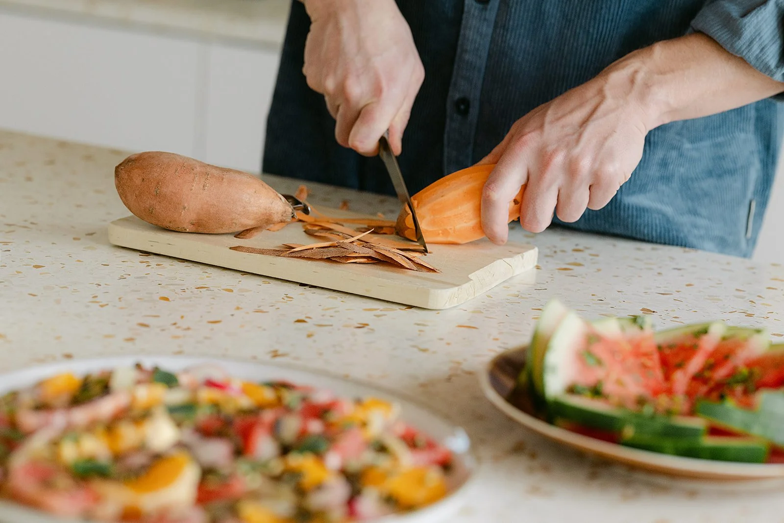 Persoon snijdt een zoete aardappel en een yam op een snijplank in een keuken, terwijl er in de voorgrond een gevarieerde salade en een schaal gerookte komkommer geserveerd worden.