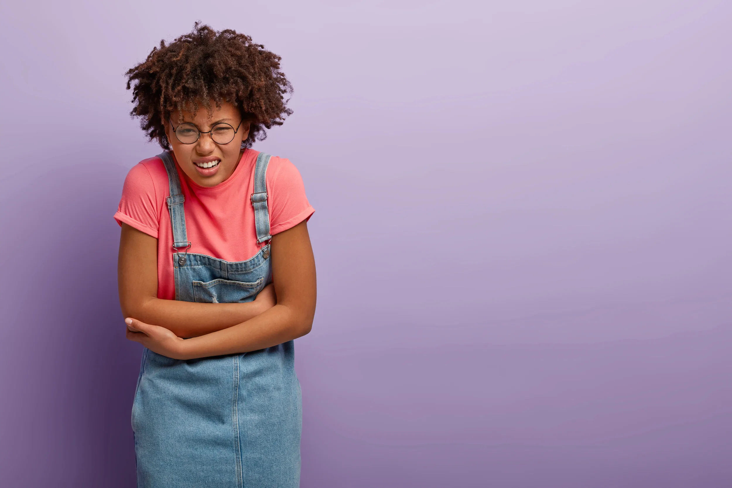 Een vrouw met een verwonde buik, gekleed in een roze shirt en een denim overall, staat tegen een lila achtergrond. Ze heeft een pijnlijke uitdrukking op haar gezicht.