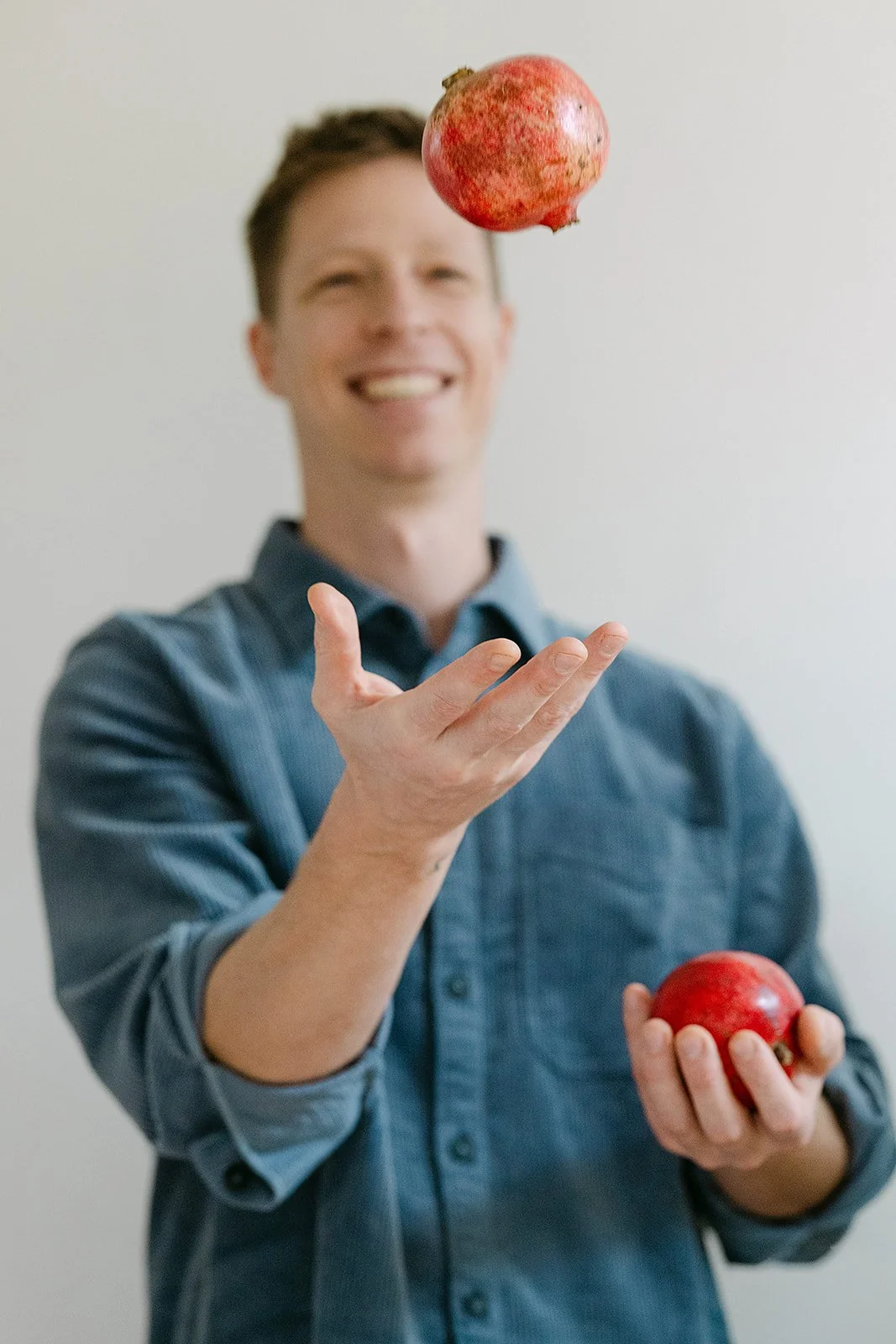 Man die één granaatappel vasthoudt en een andere appel in de lucht gooit, lachend en lachend, terwijl hij een blauwe overhemd draagt.