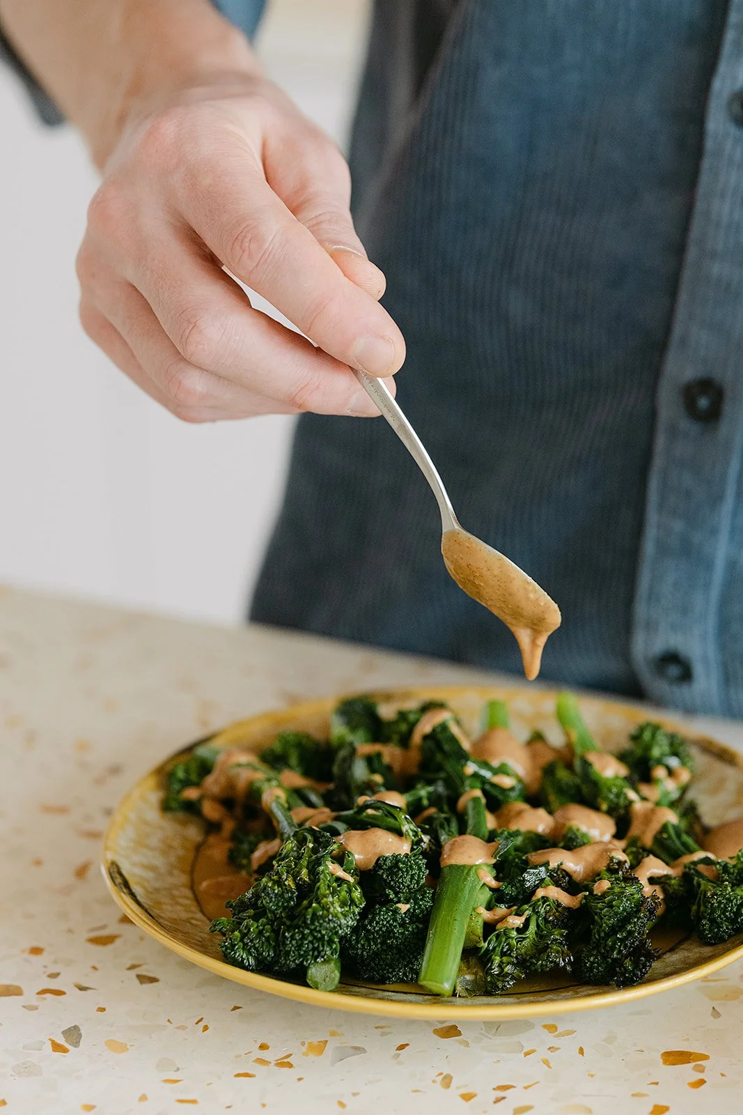 Een hand die een lepel met een saus of dressing boven een bord met gestoomde broccoli bedekt