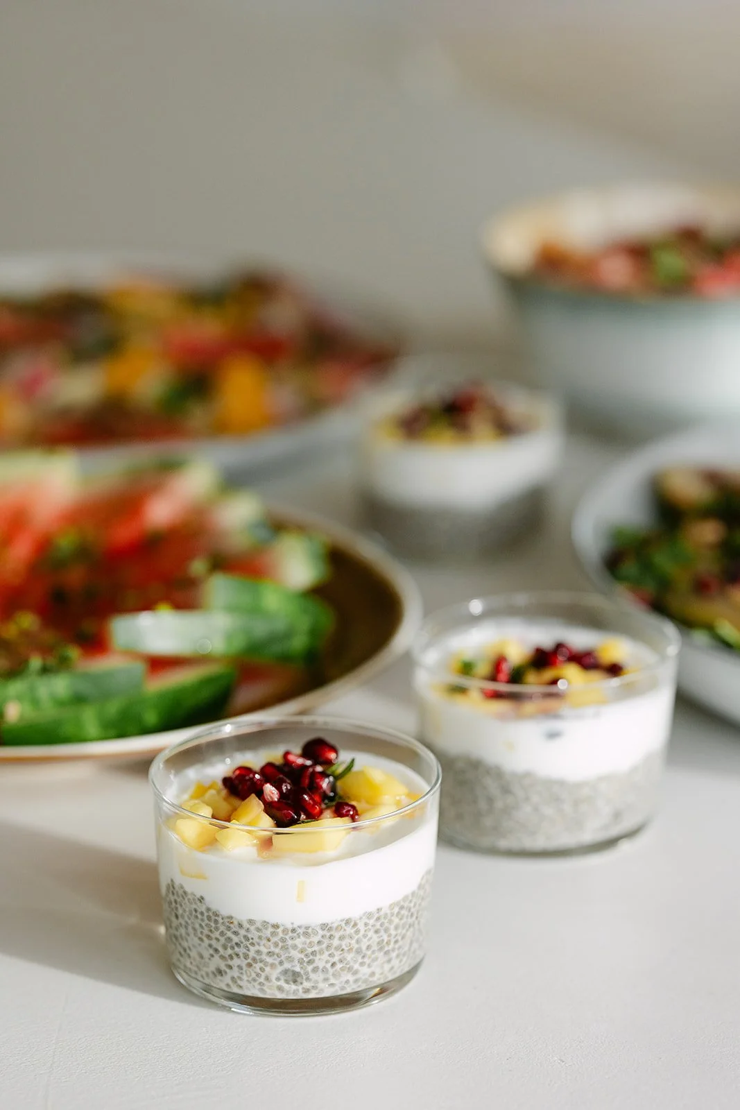 Twee kleine dessertglaasjes met chiazaadpudding, versierd met fruit en granola, op een witte tafel met verschillende schalen eten op de achtergrond.