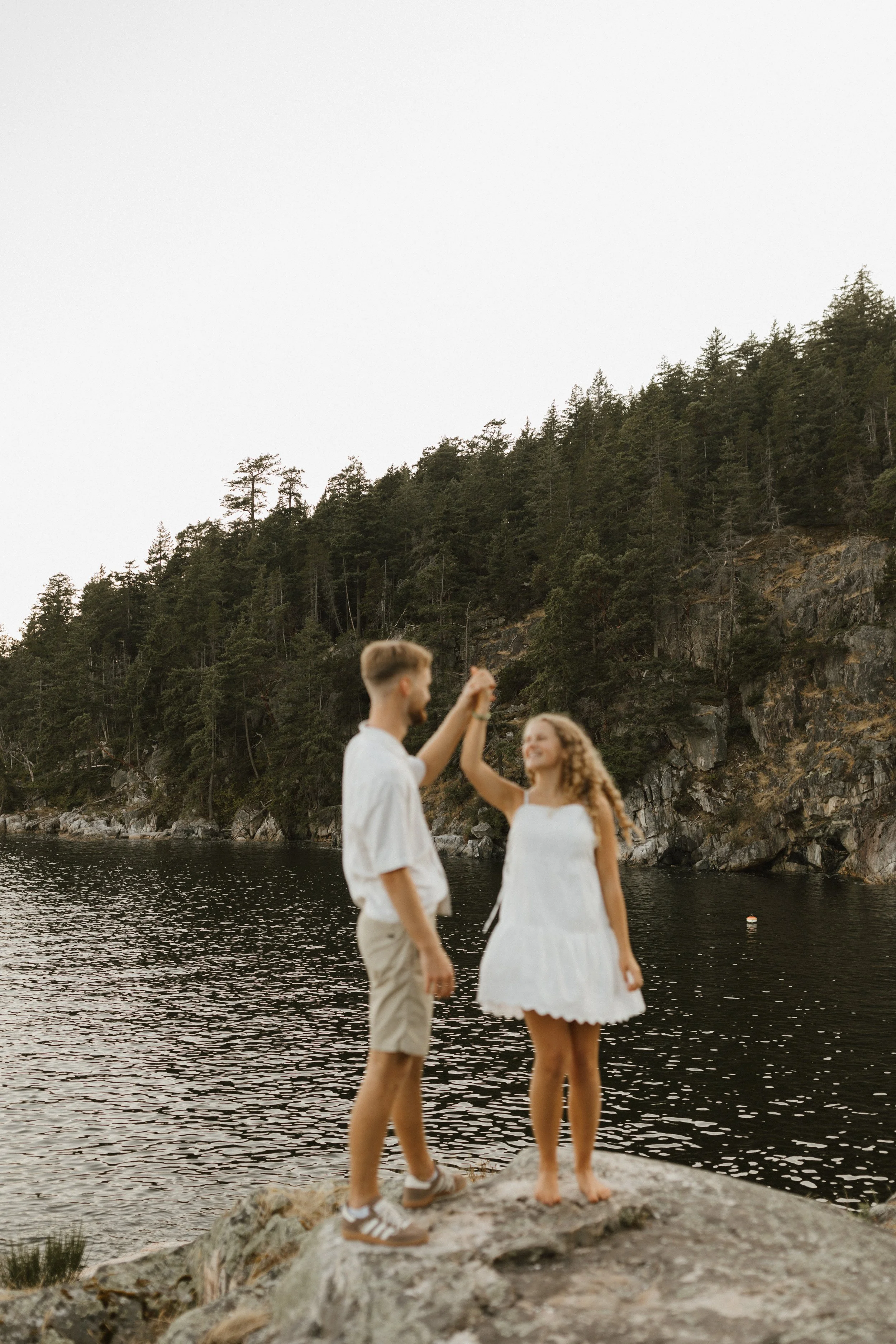 A young couple dancing on a rock by a lake with a forested hillside in the background.