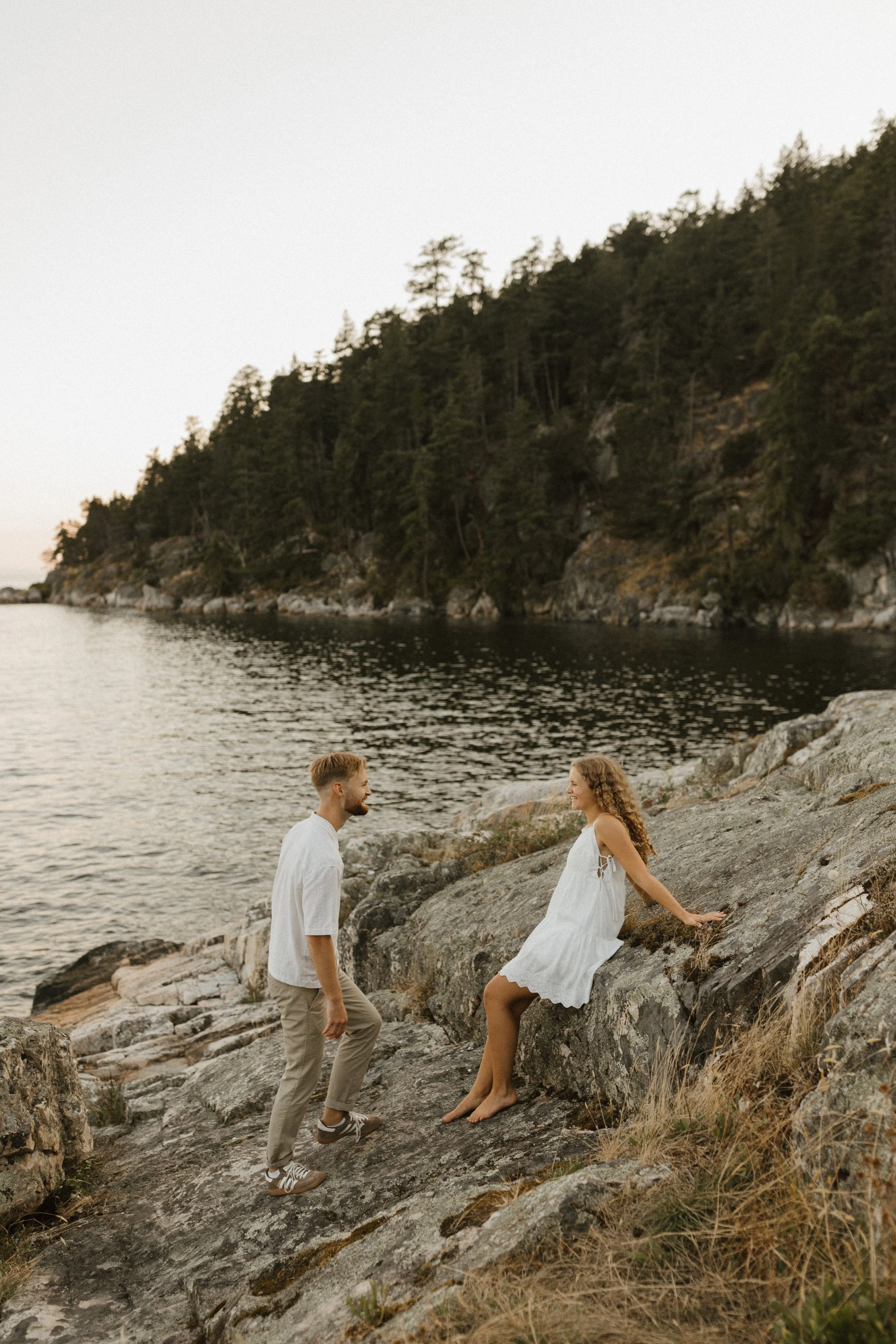 A young man and woman are on a rocky shoreline near a body of water, with a forested hillside in the background. The woman, sitting on a large rock, wears a white dress and has curly hair. The man, standing in front of her, wears a white shirt and be