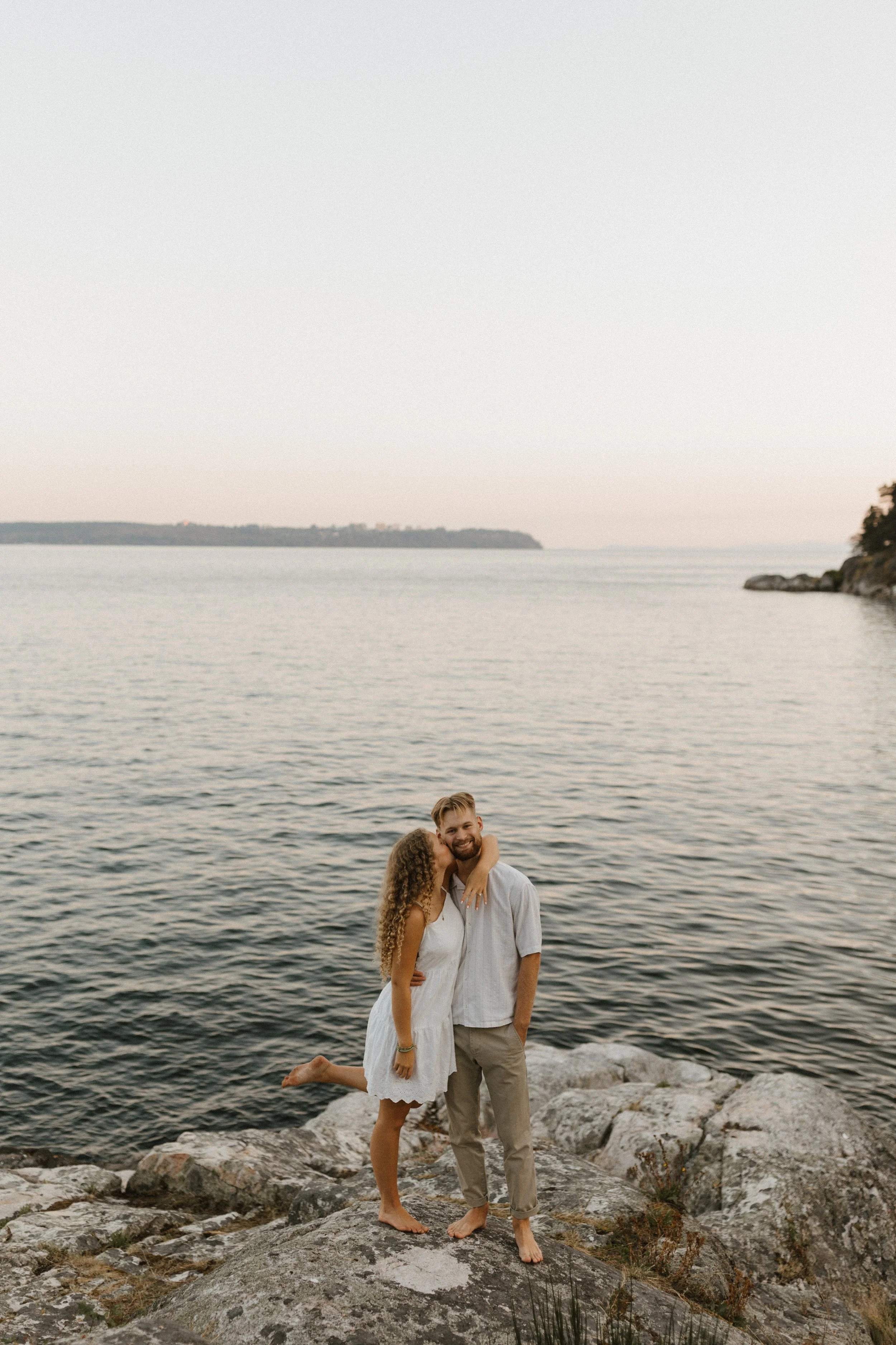 A couple standing on rocks by the water at sunset, with the woman kissing the man's cheek as he smiles.
