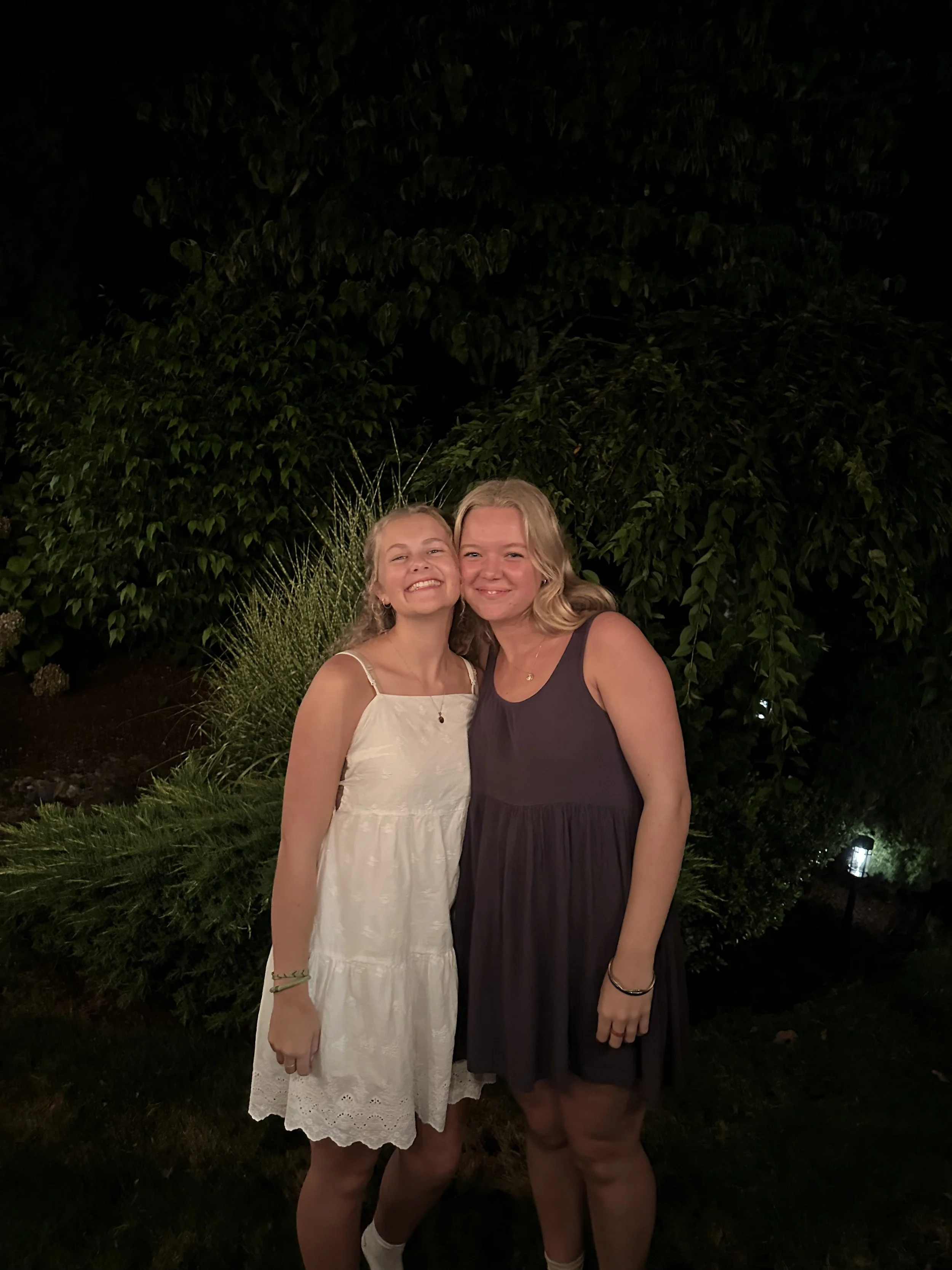 Two young women smiling and posing outside at night, standing close together in front of greenery.