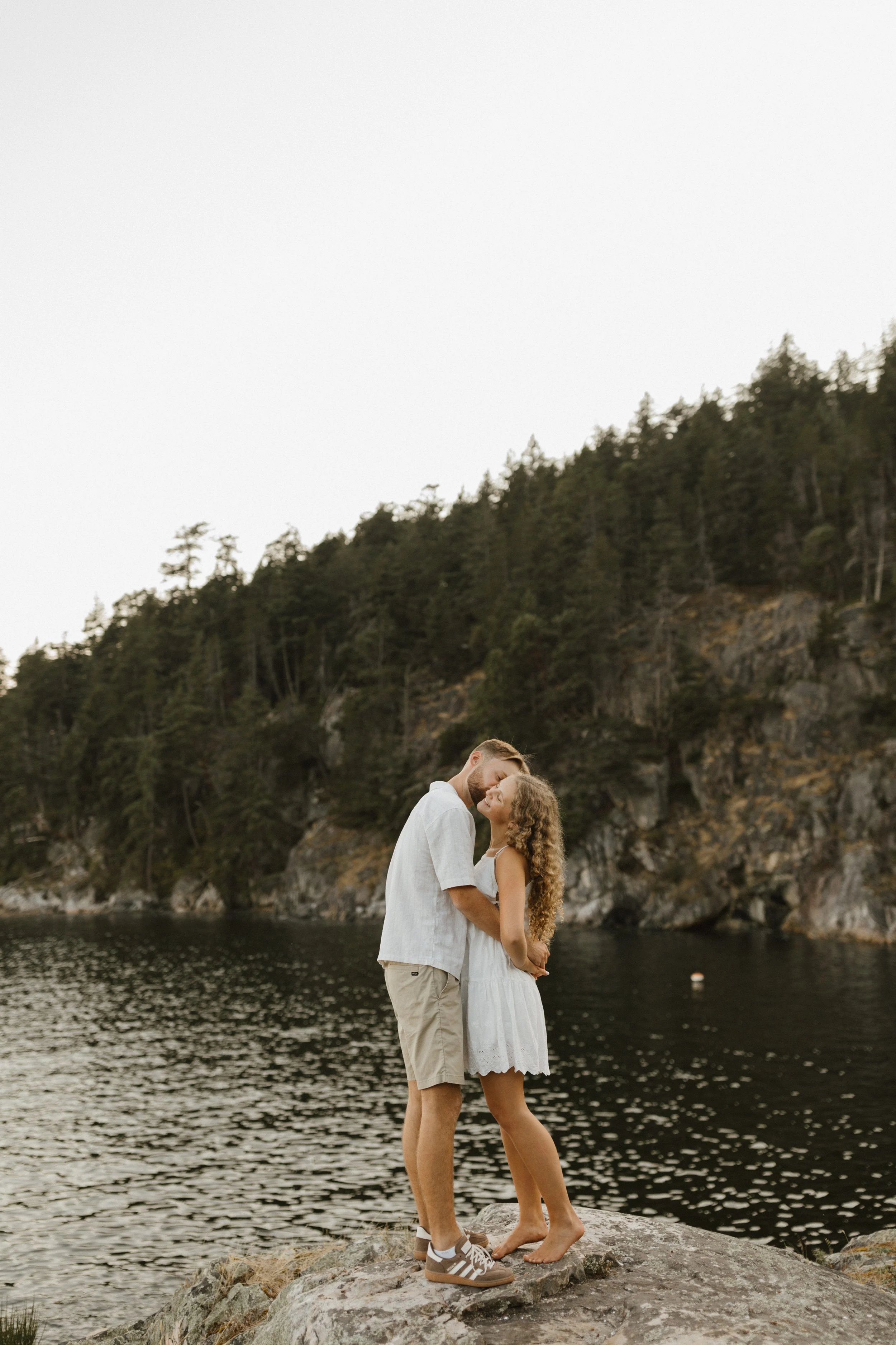 A couple standing on a rock by a lake, embracing and touching foreheads, with forested hills in the background.