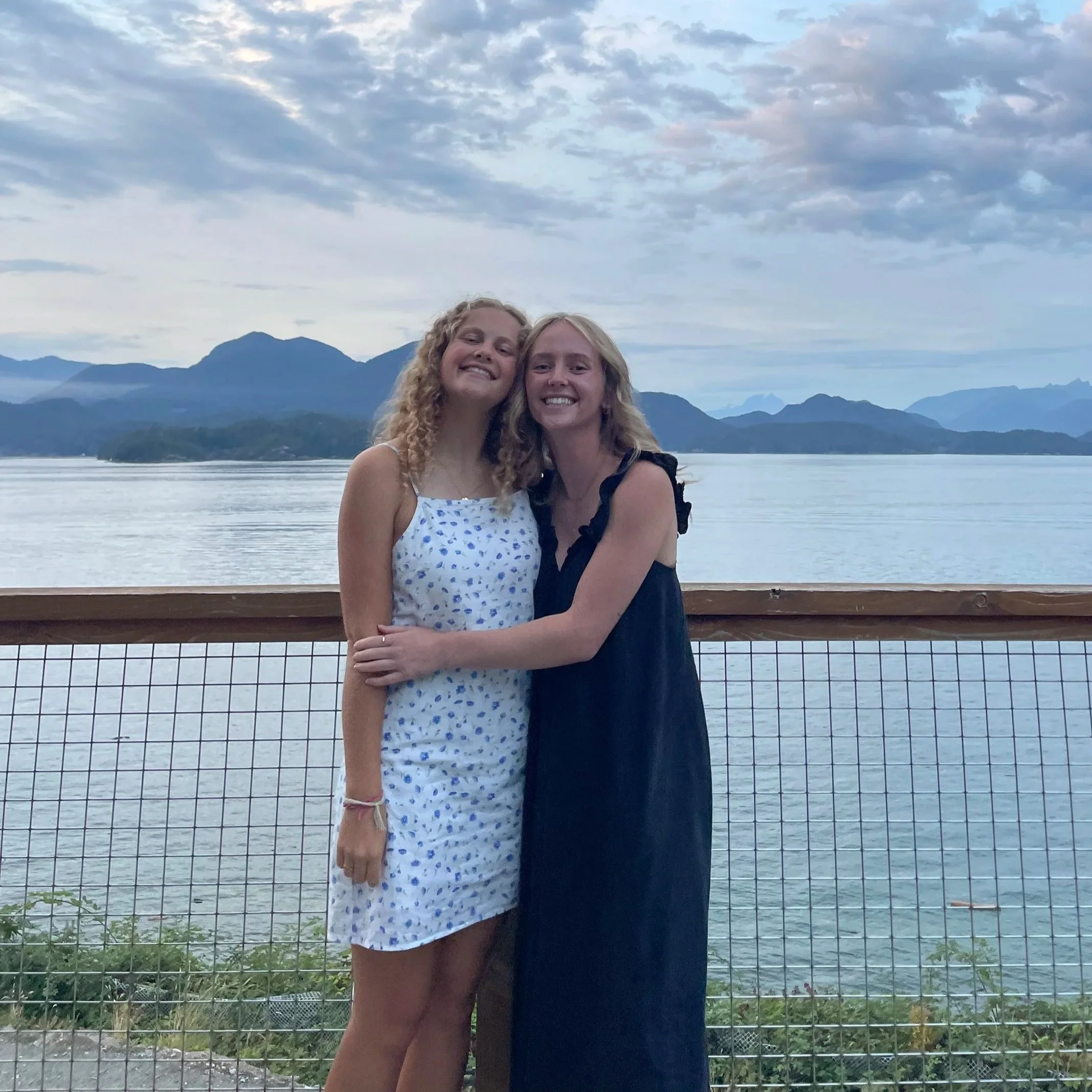 Two young women hugging and smiling at the camera near a lake, with mountains and a cloudy sky in the background.