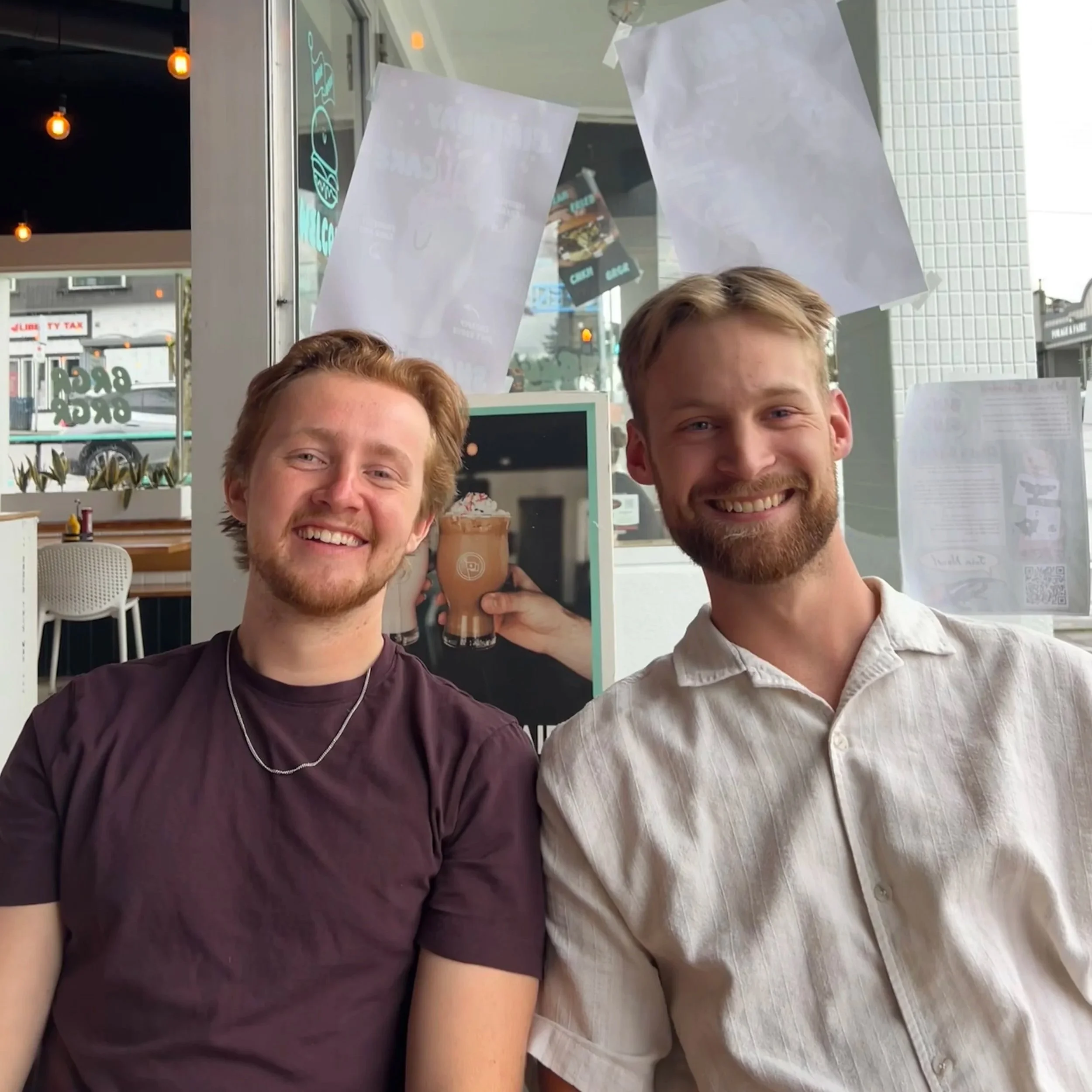 Two young men with beards smiling and sitting together inside a cafe. The man on the left has red hair and a maroon shirt, and the man on the right has light brown hair and a light-colored shirt. There are windows and posters behind them, and a blurred outside view of buildings and cars.
