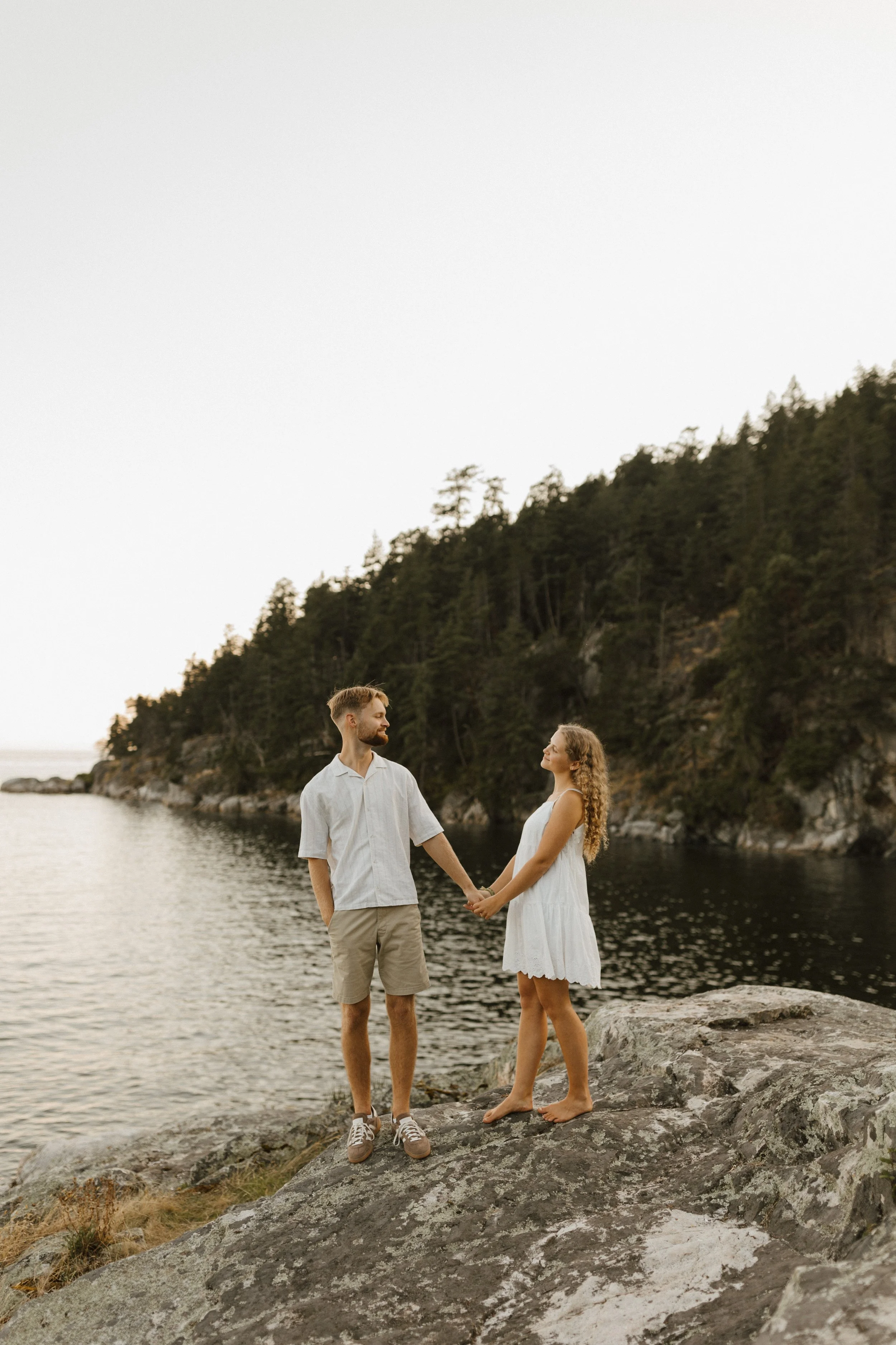 A man and woman holding hands on a rocky shoreline near water, with a forested hillside in the background during sunset.