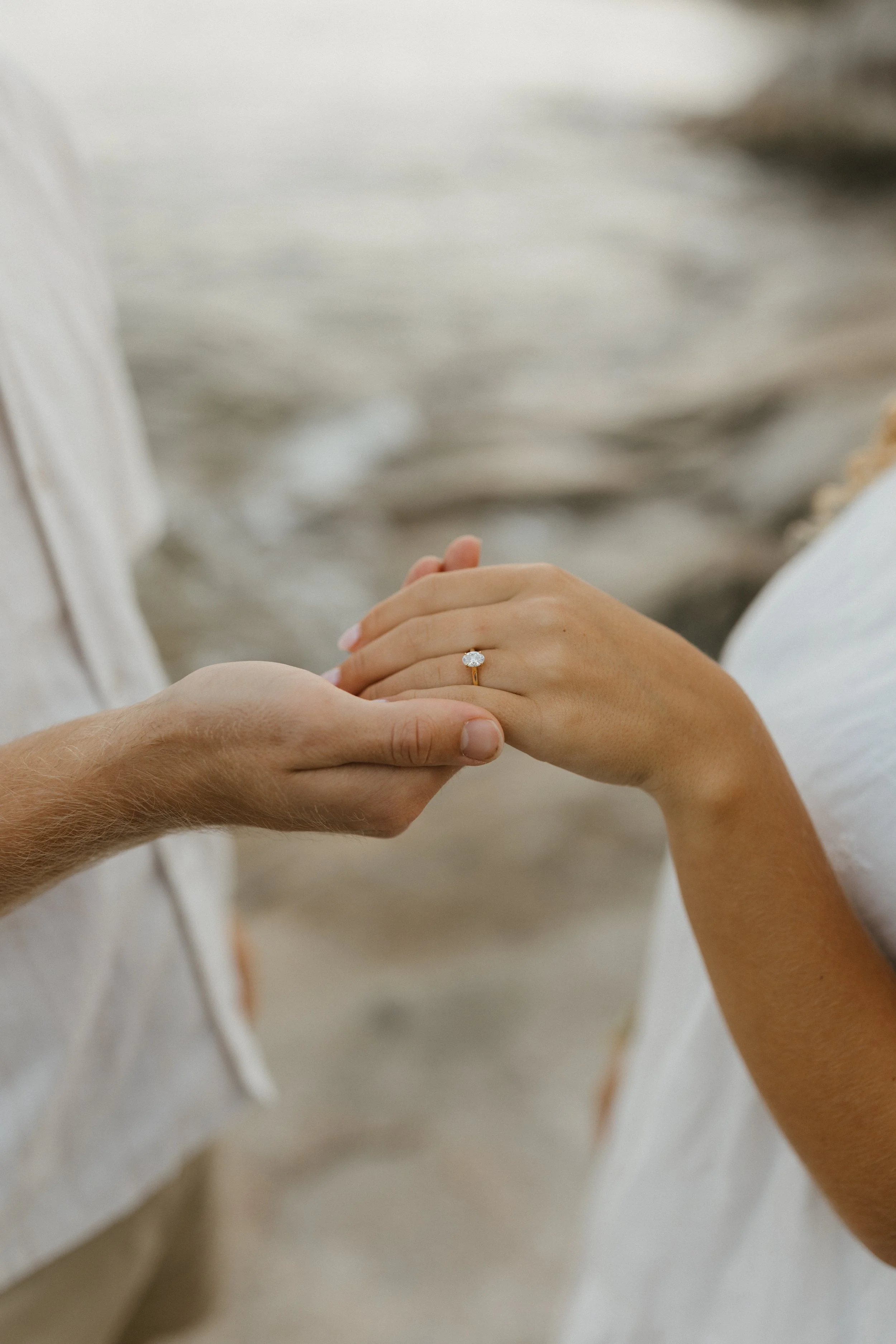 Close-up of a couple holding hands, with the woman's hand showing an engagement ring, on a beach.