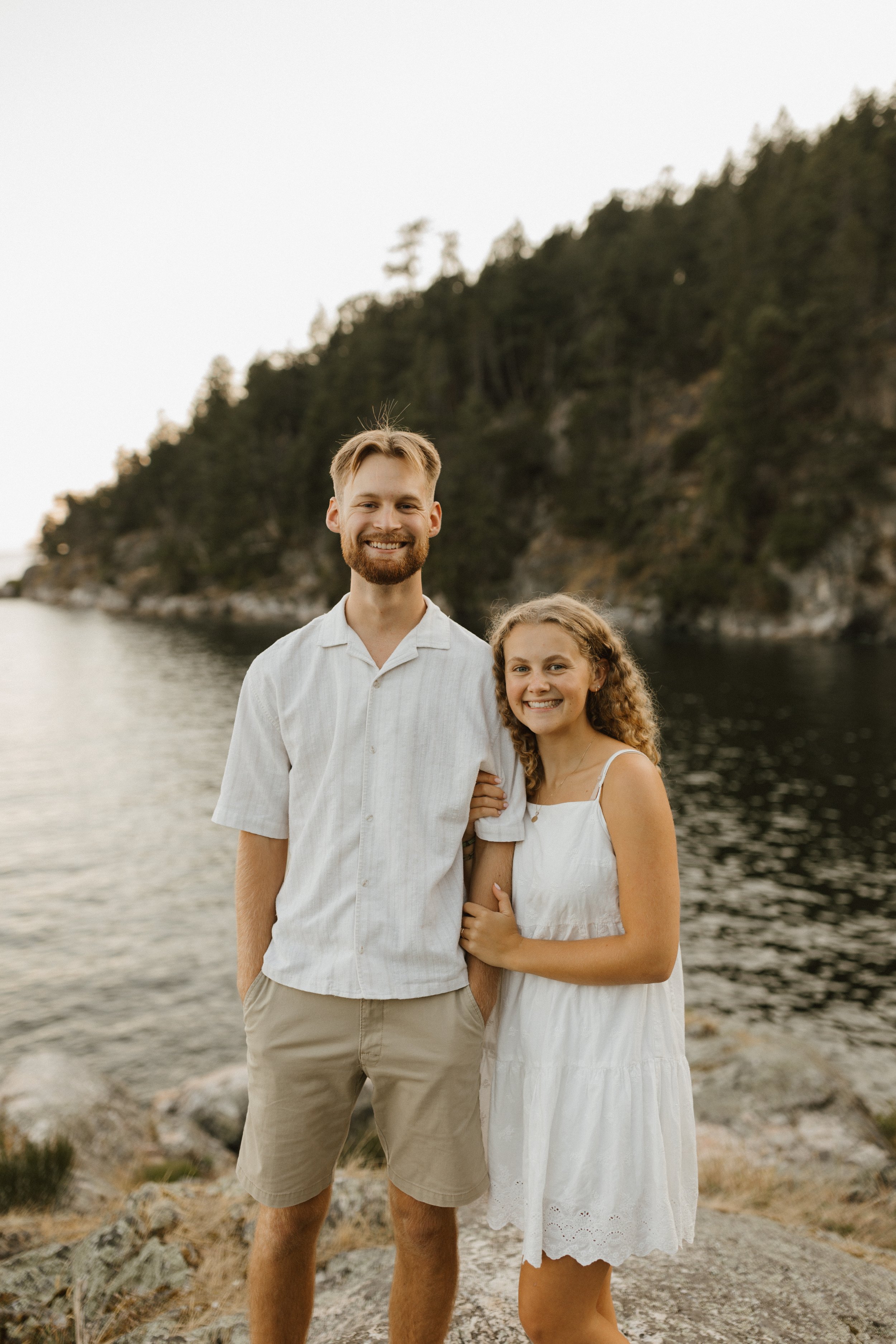 A smiling couple standing arm-in-arm near a body of water with a forested hillside in the background.