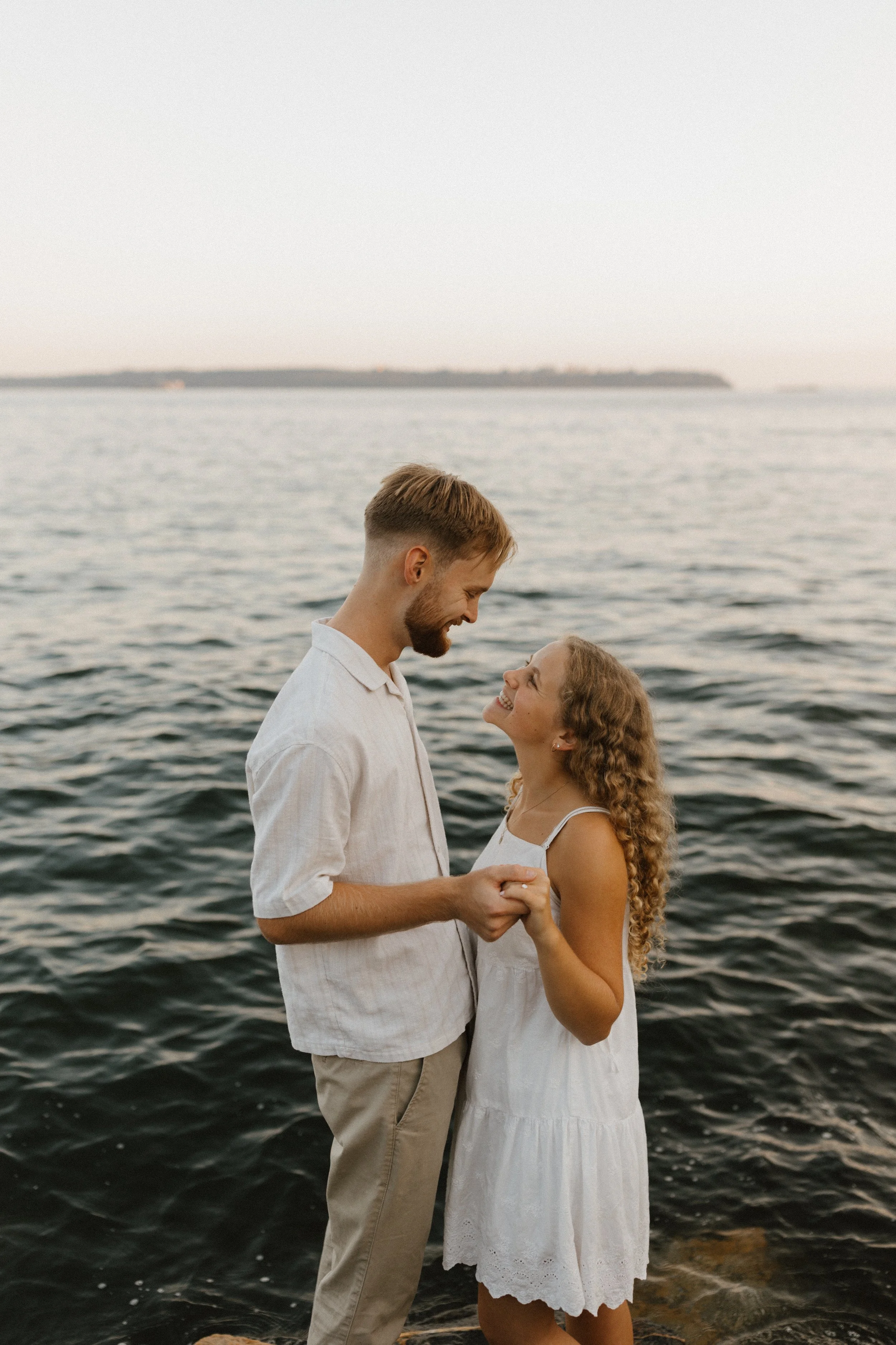 A smiling couple holding hands in front of a body of water, with a distant shoreline in the background.