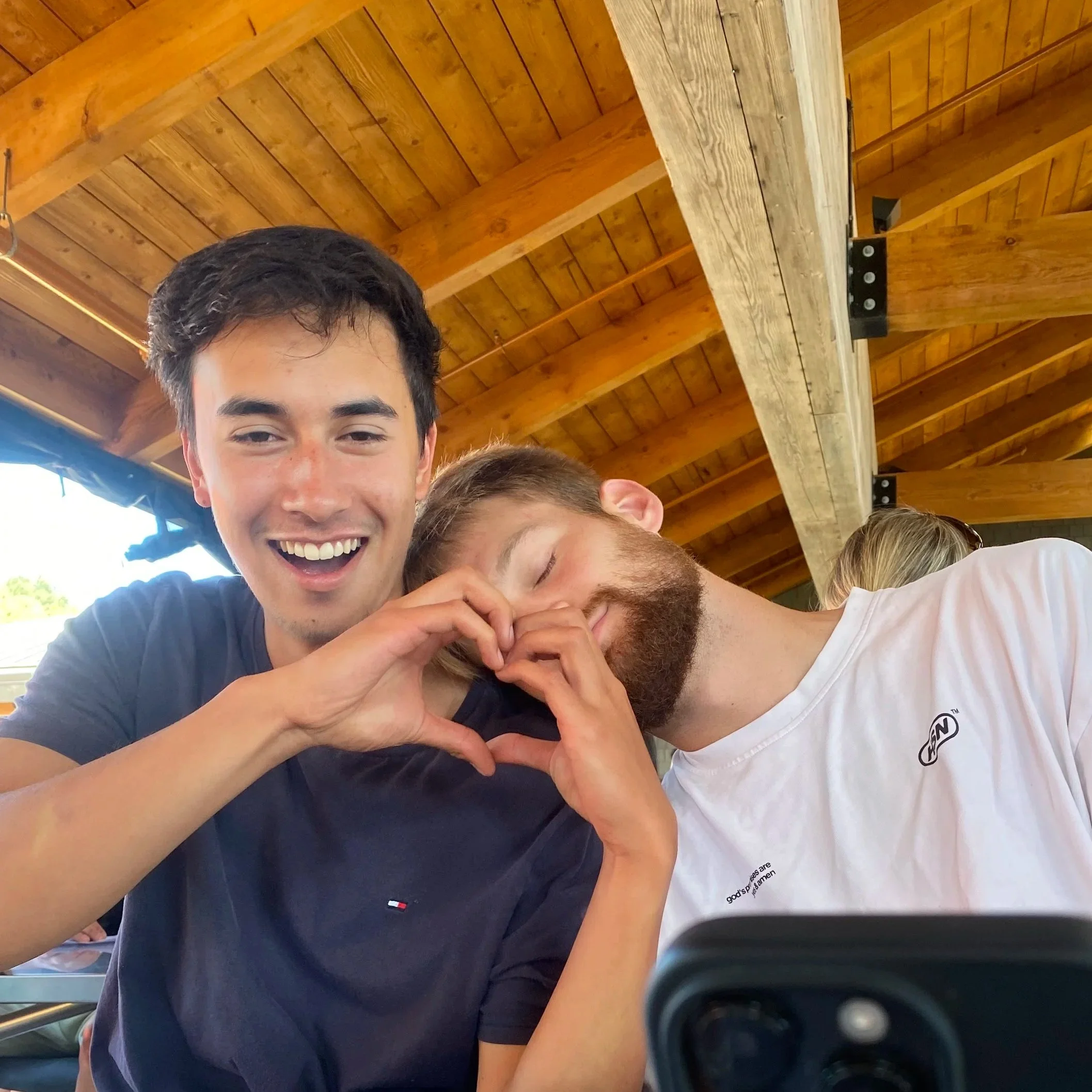 Two young men smiling and forming a heart shape with their hands while sitting under a wooden roof. One is wearing a navy shirt, and the other has a white shirt with a logo. They appear happy and are posing for a photo.