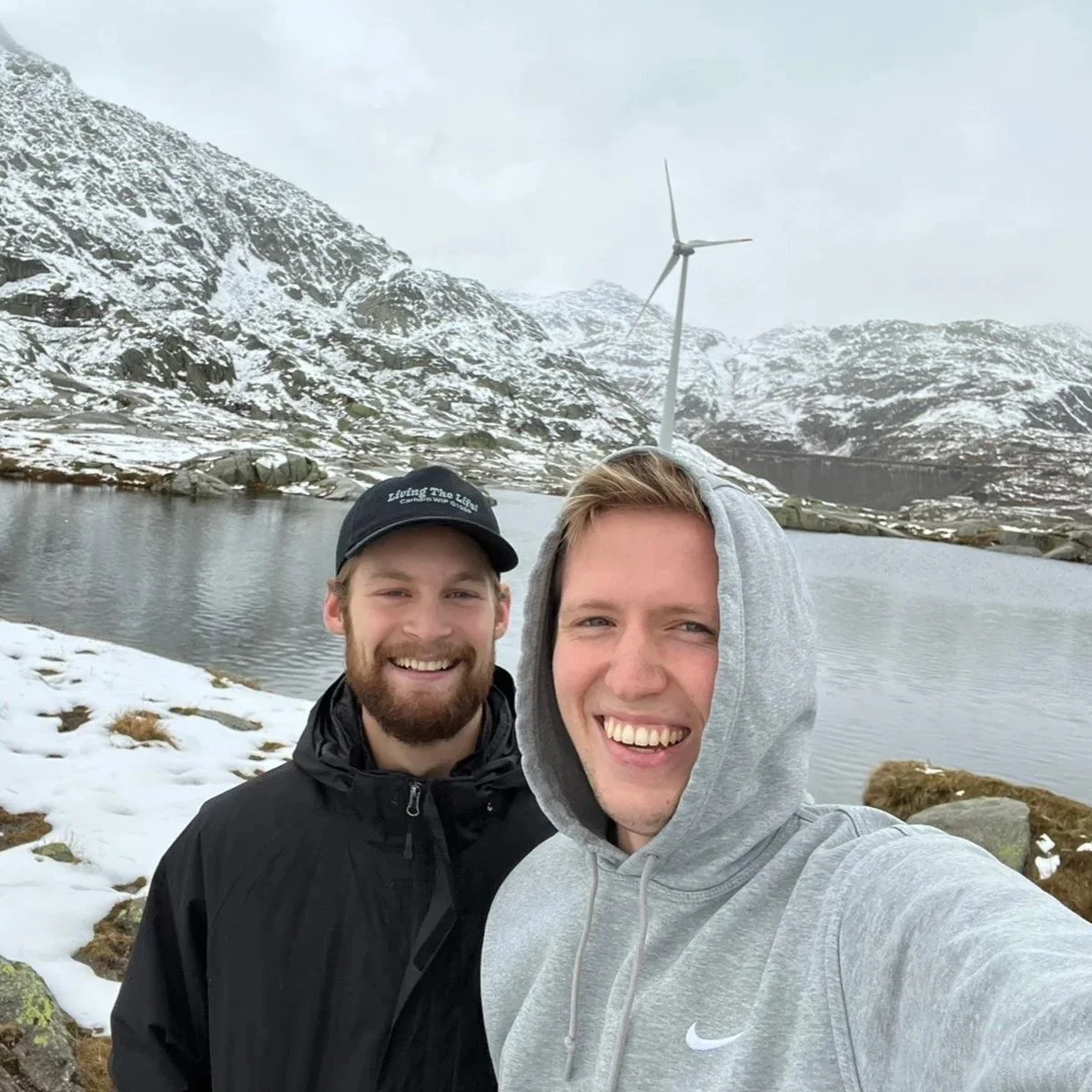 Two smiling men taking a selfie in a snowy mountainous landscape with a lake, rocks, and a wind turbine in the background.