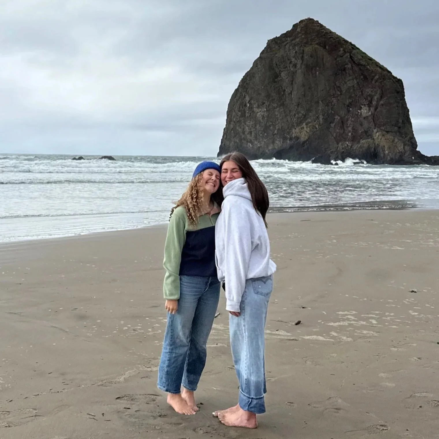Two women are standing on a sandy beach, smiling and hugging each other, with a large rock formation and ocean waves in the background on an overcast day.