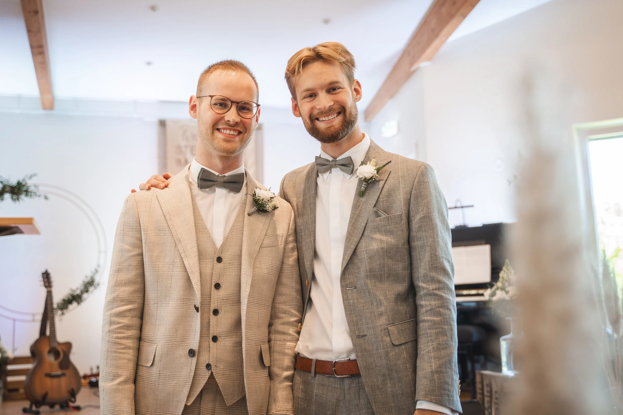 Two men wearing suits and bow ties standing inside a venue, smiling, with floral boutonnieres on their lapels.