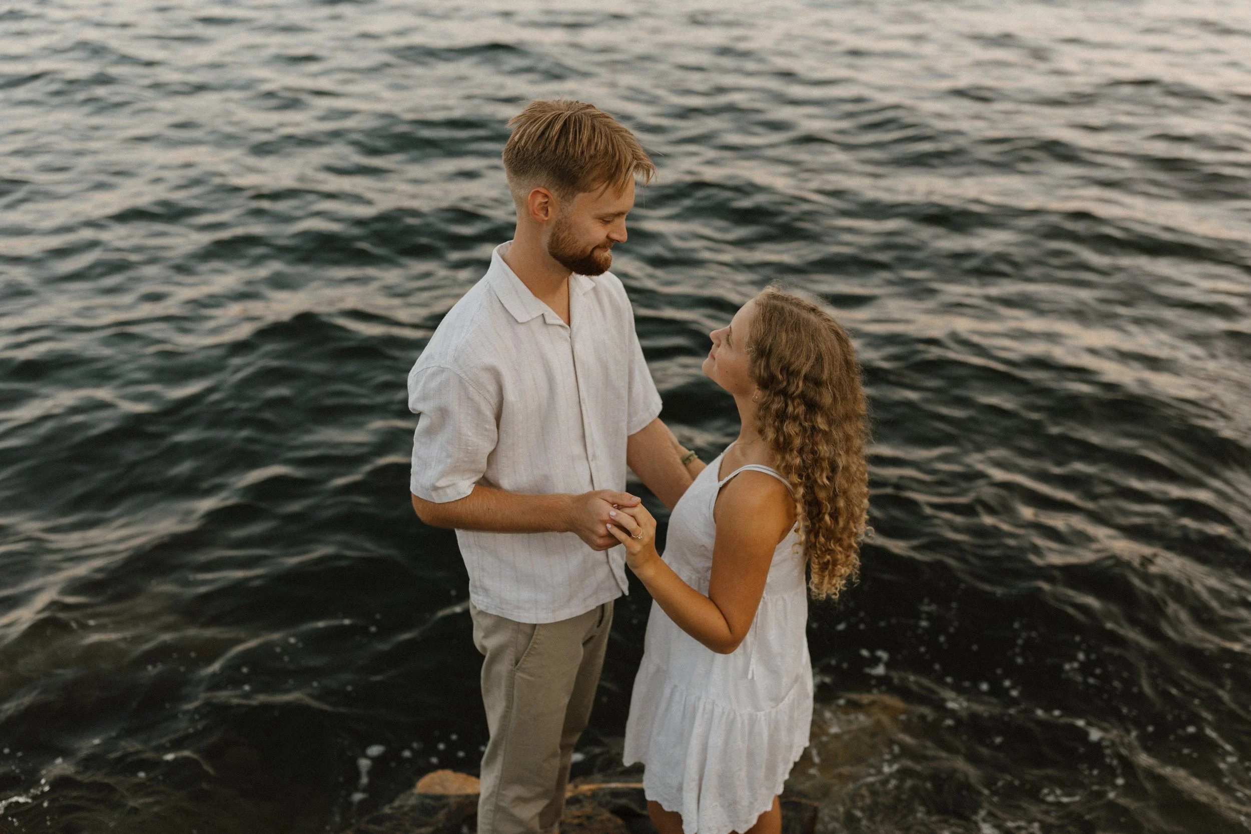 A young couple stands close together on rocks by the water, holding hands and looking into each other's eyes during sunset.