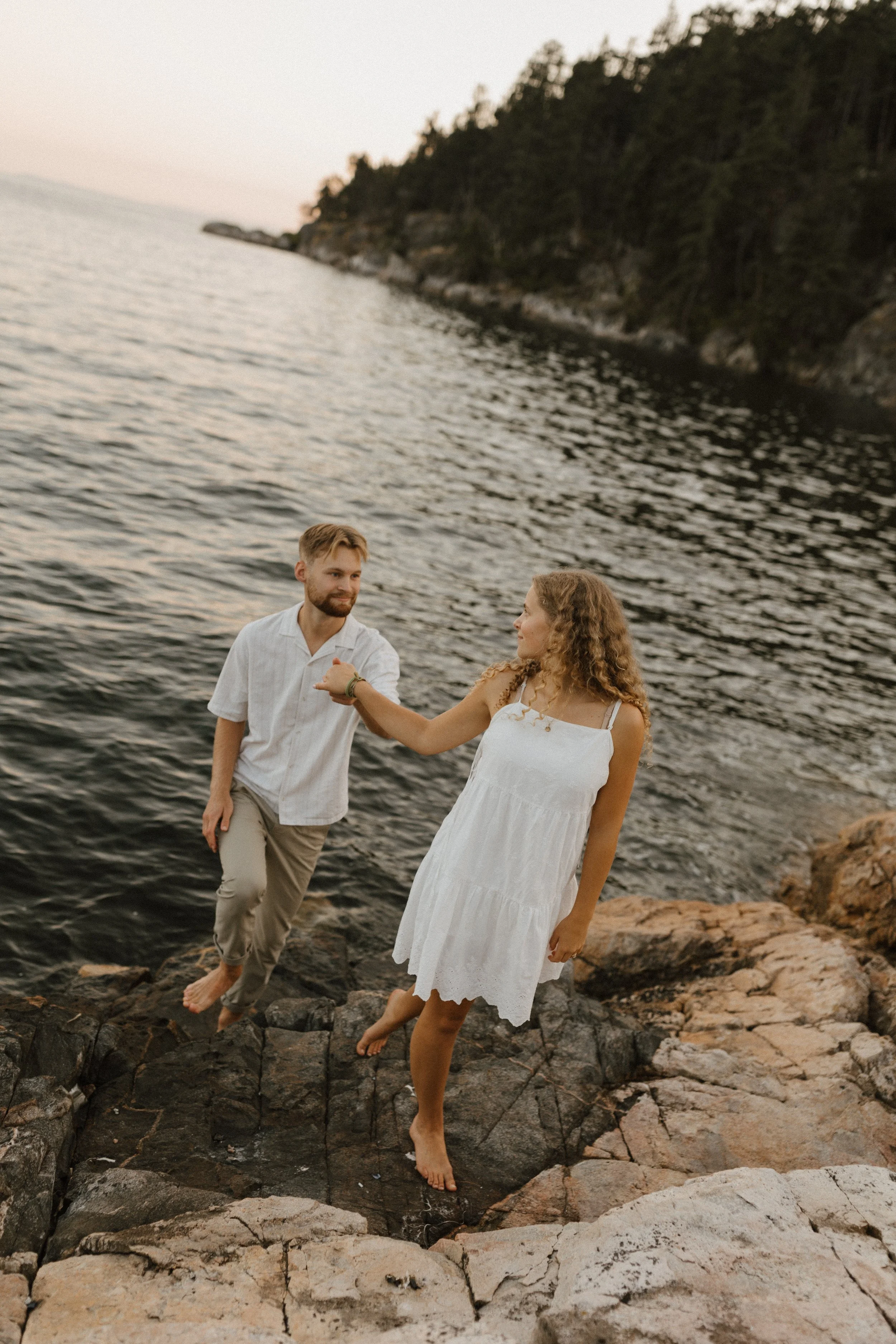 A young couple dressed in white attire playfully balancing on rocks by a body of water at sunset.