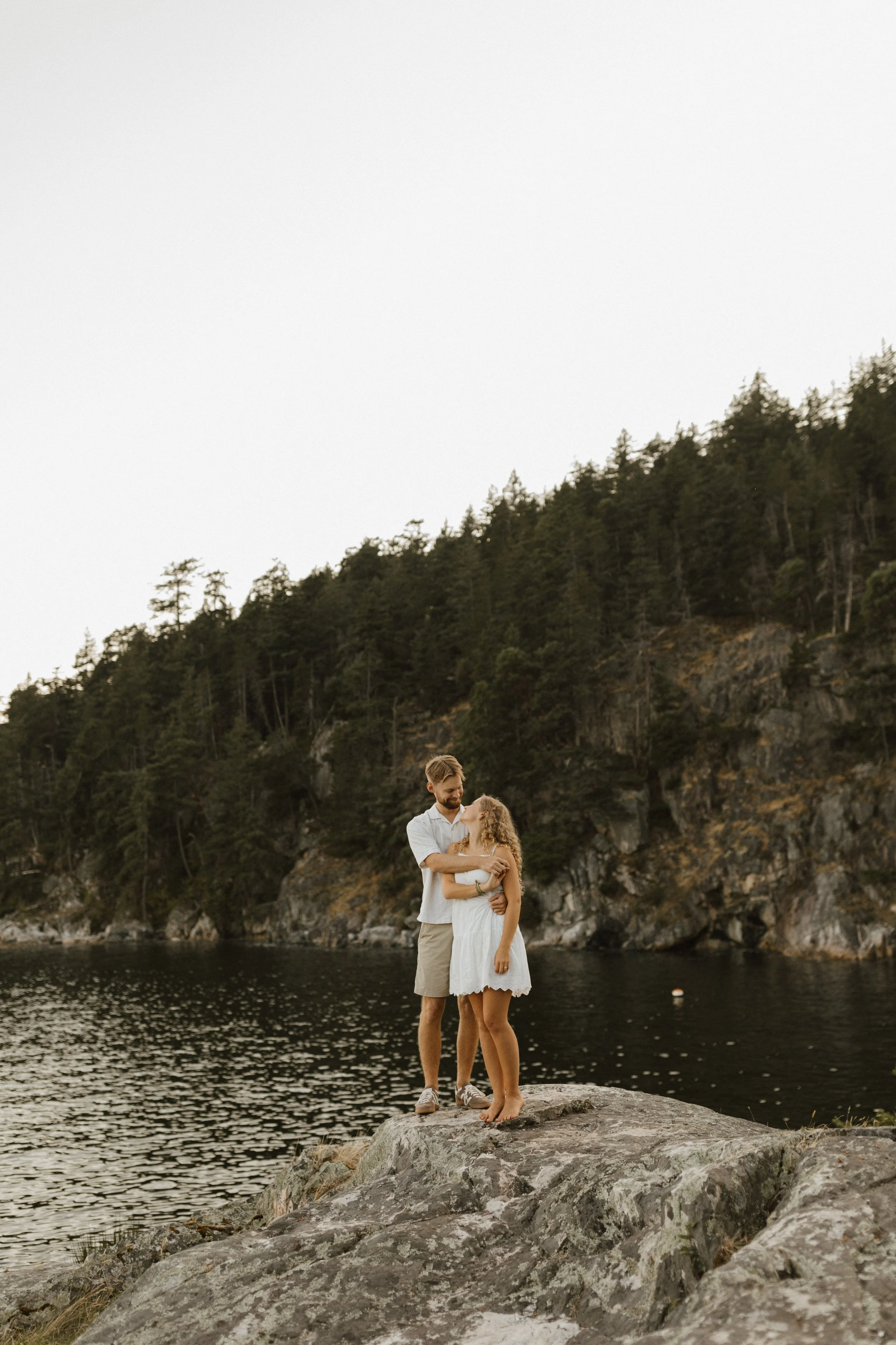 A couple standing on a rock near a body of water, with a forested hillside in the background, embracing each other during sunset.