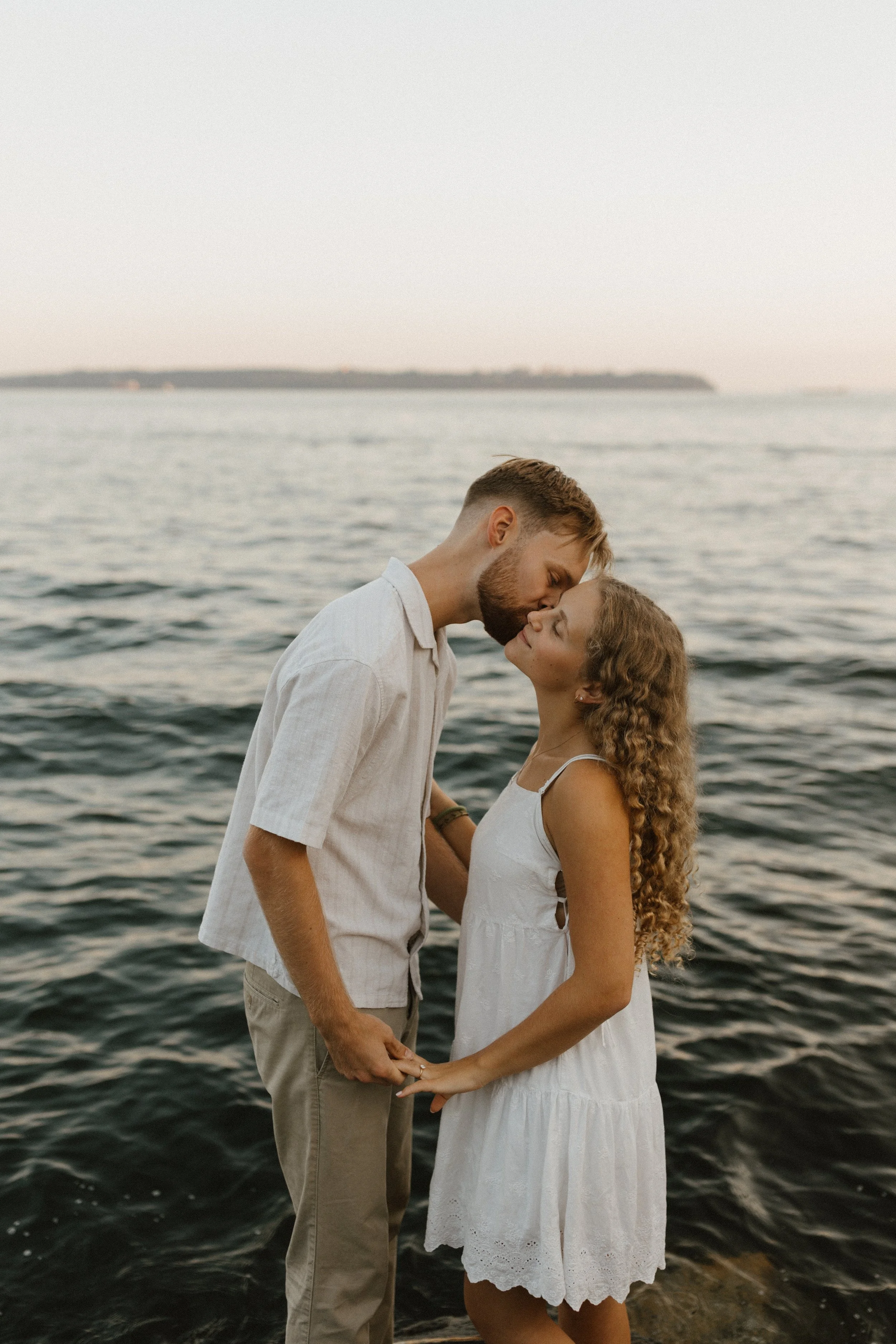 A couple in white clothing sharing an intimate moment by the water, with the man about to kiss the woman on her forehead, during sunset or sunrise.