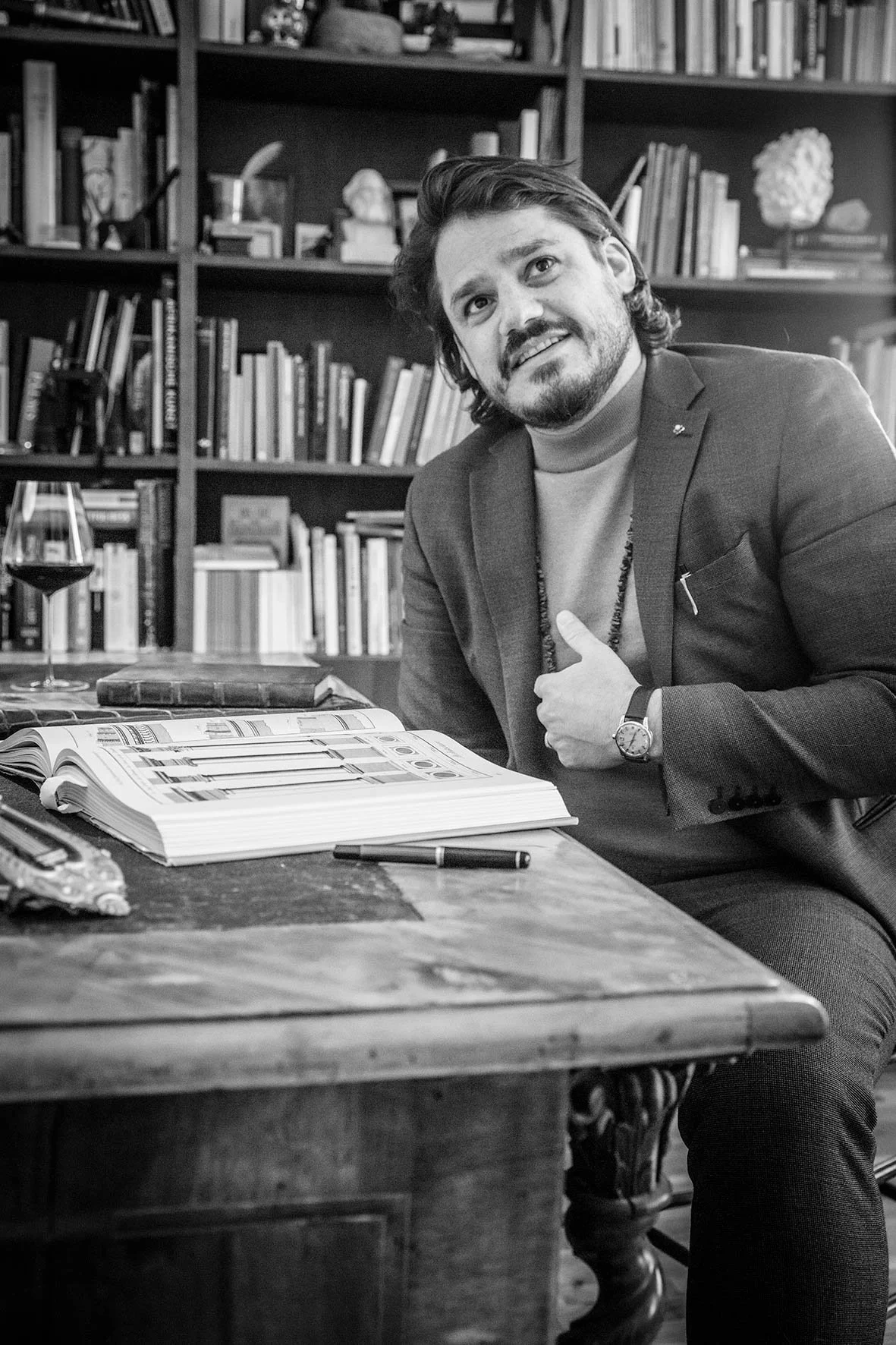 Mann sitzt an einem großen Holztisch mit einer offenen Buch- und einer Zeitschriftenseite, eine Weinflasche und ein Glas Wein auf dem Tisch, im Hintergrund Bücherregale.