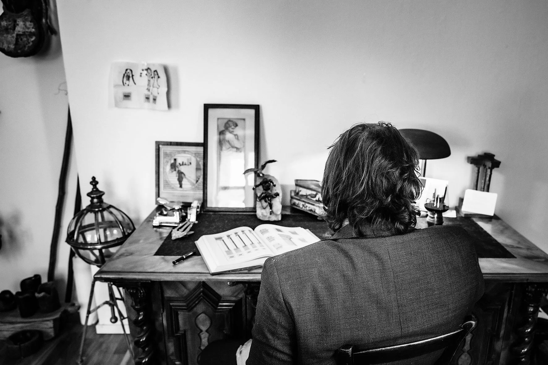 Eine Person sitzt an einem Schreibtisch, liest ein Buch, mit Bildern und Dekorationen auf dem Tisch und an der Wand, schwarz-weiß.