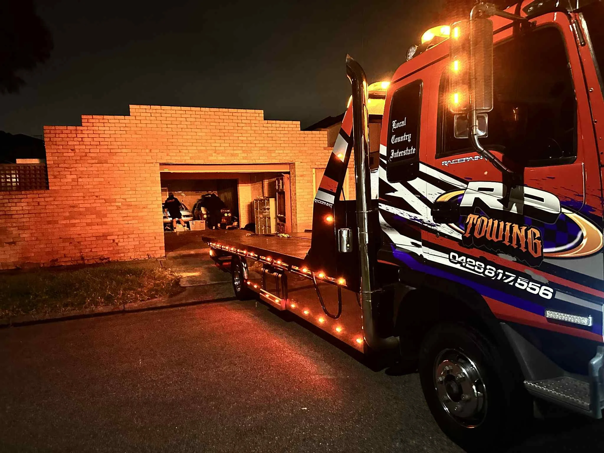 Night scene of a tow truck parked in front of a red brick structure with an open garage. Inside the garage, two people are working near vehicles.