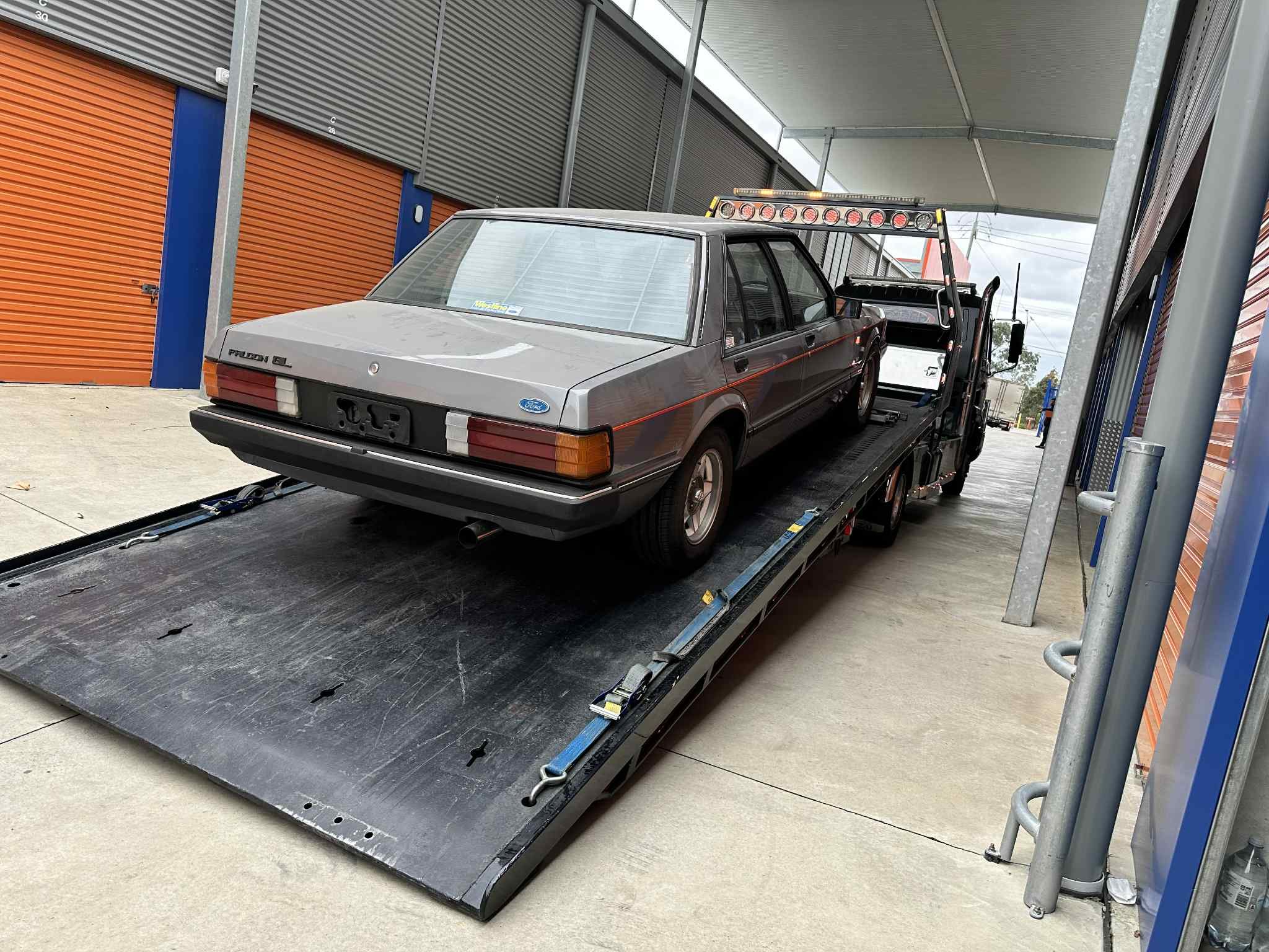 Gray vintage Ford Pinto GL on a flatbed tow truck under a canopy at a storage or service facility.