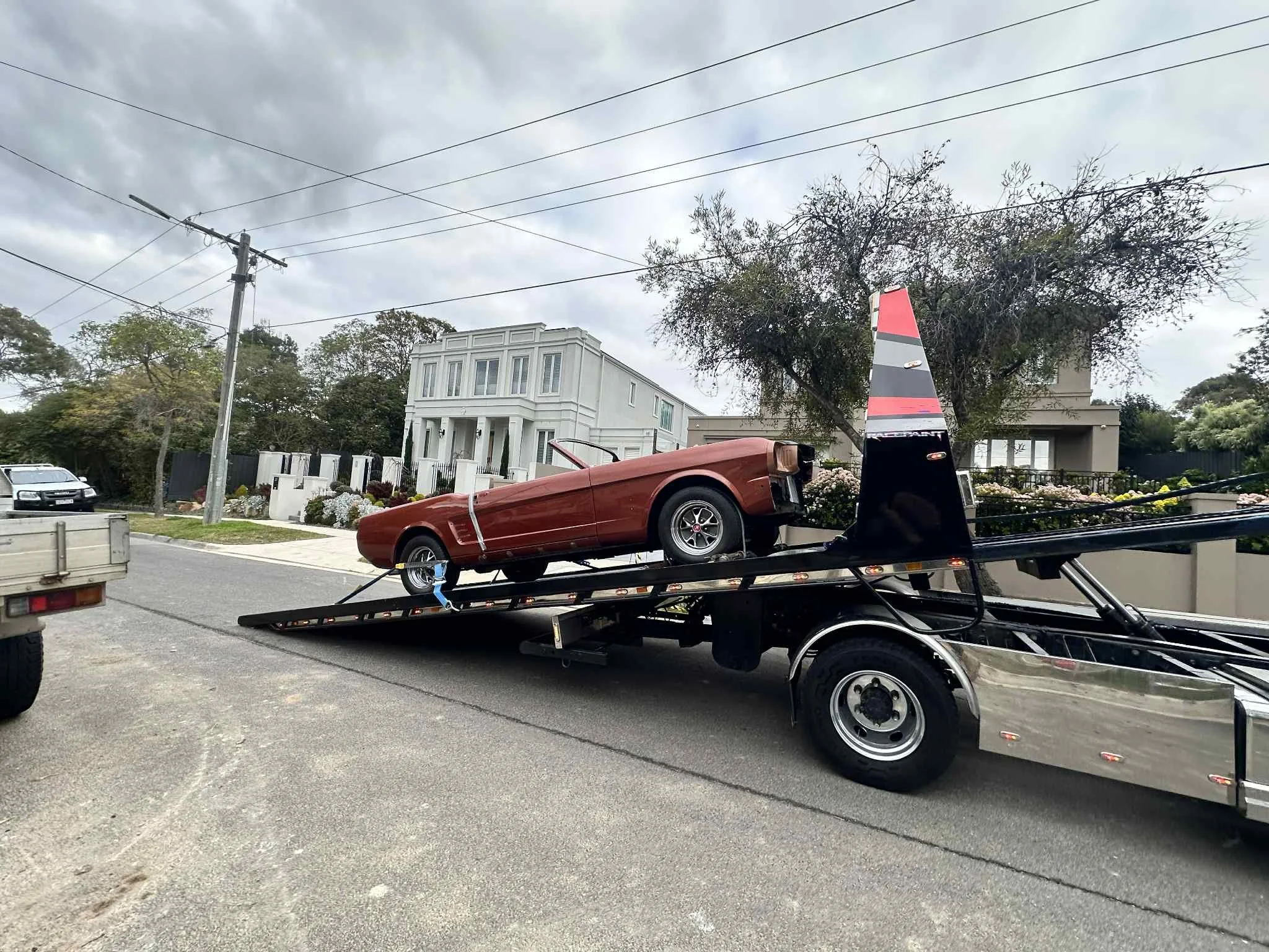 A vintage red convertible car on a flatbed tow truck parked on a residential street with modern houses, trees, and power lines in the background.