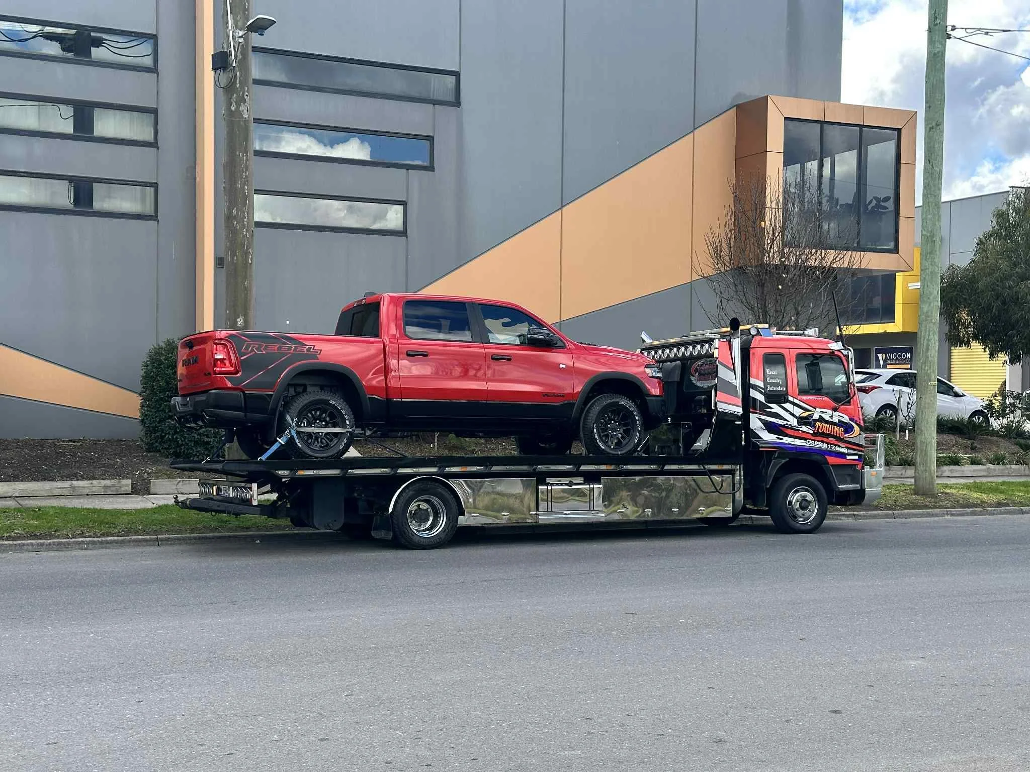 Flatbed tow truck carrying a red RAM Rebel pickup truck in front of a modern building with angular design and large windows, on a city street.