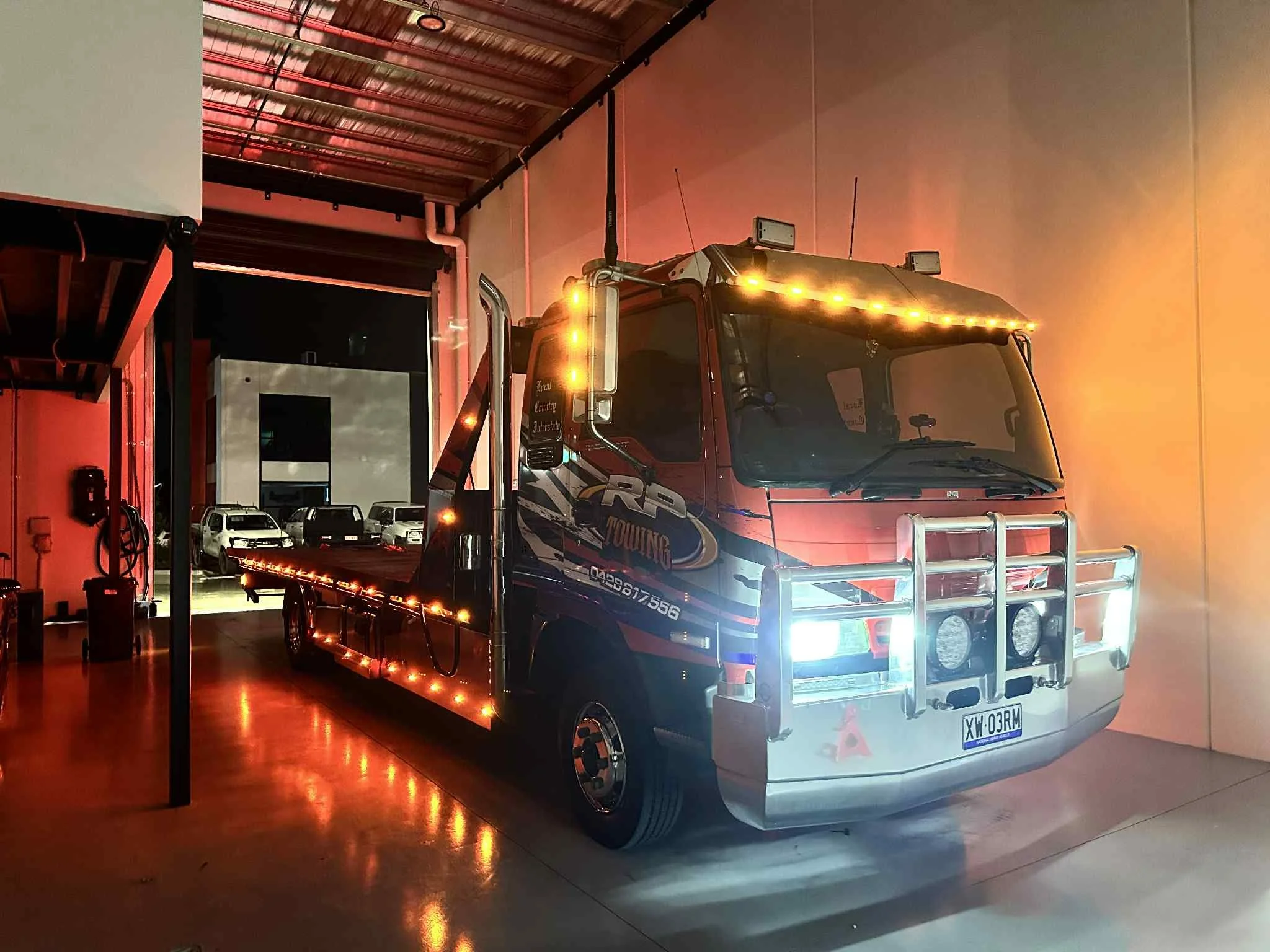 A flatbed tow truck with bright lights parked inside a garage, with several cars visible outside through an open door in the background.