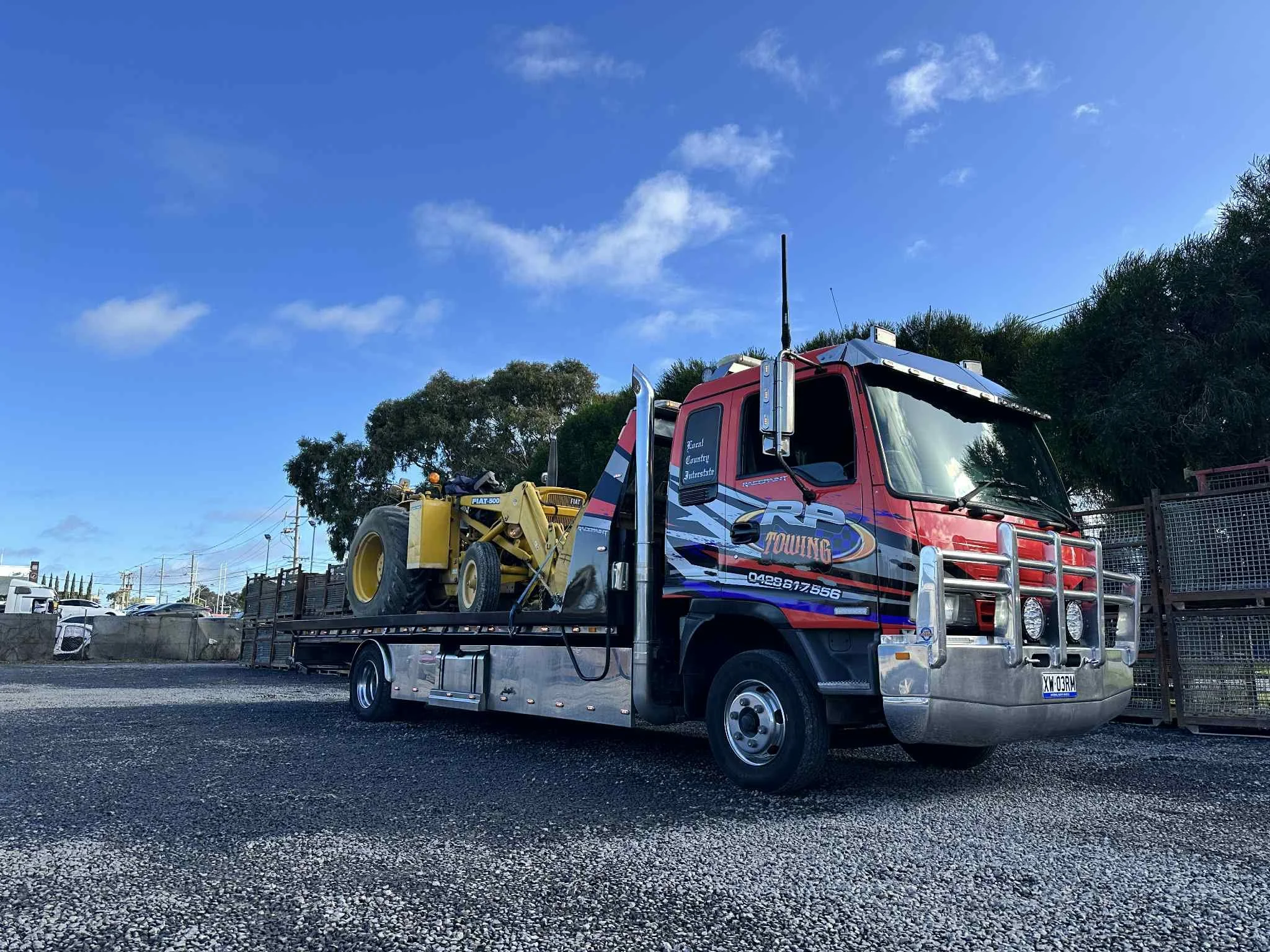A red tow truck carrying a yellow tractor on its flatbed, parked on a gravel lot with trees and a chain-link fence in the background, under a blue sky with a few clouds.