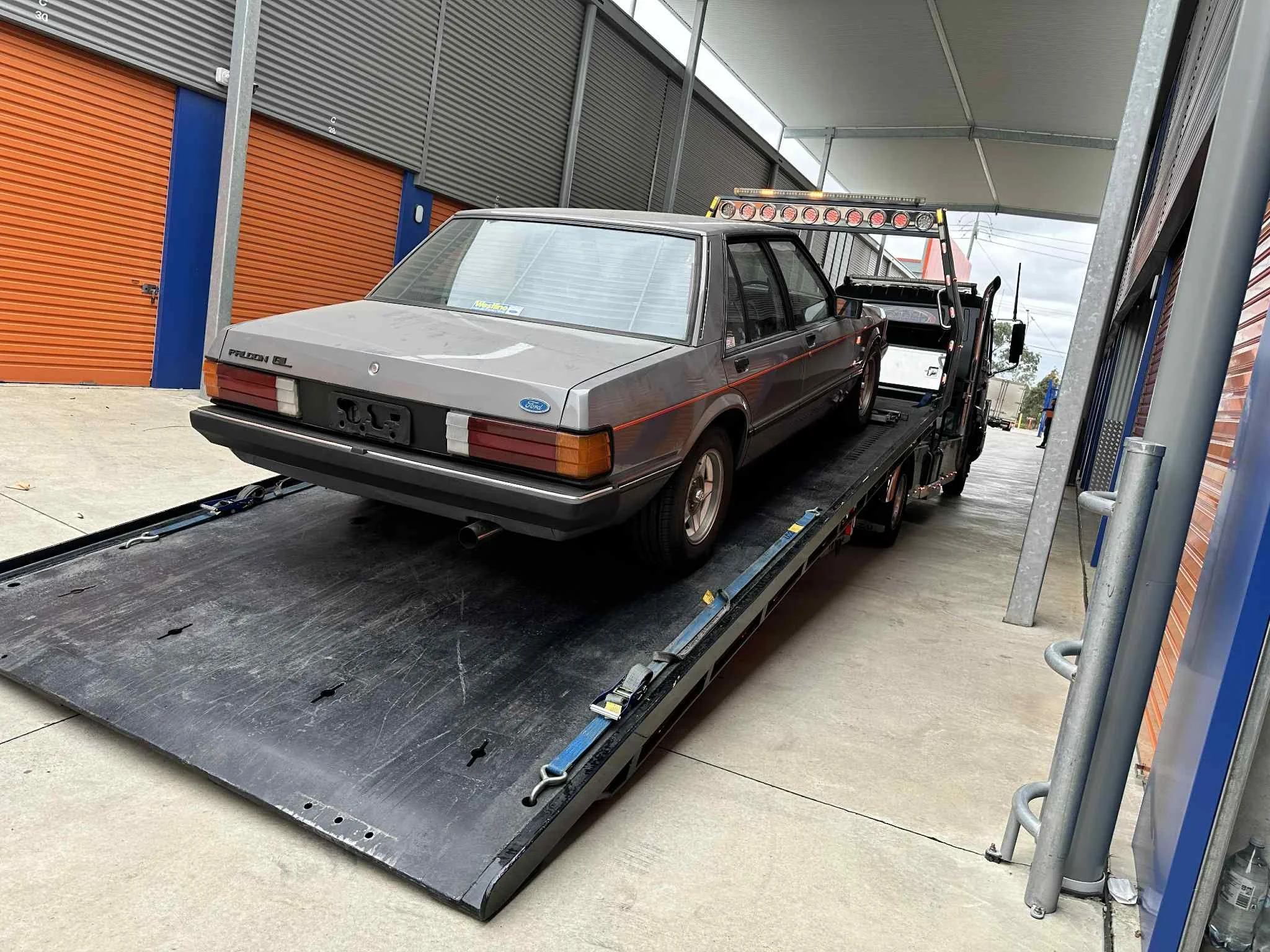Gray vintage Ford Falcon on a flatbed tow truck in a storage facility with orange and blue garage doors.
