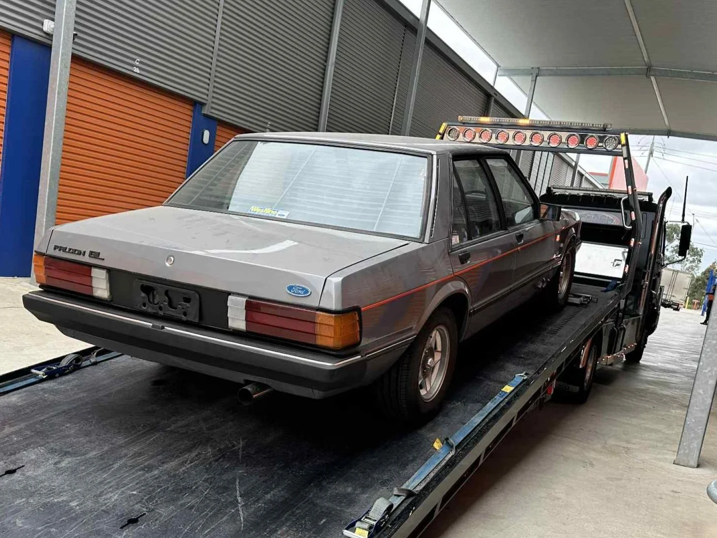 A gray Ford Falcon EL car is loaded onto a flatbed tow truck in a covered area, possibly a garage or dealership lot.